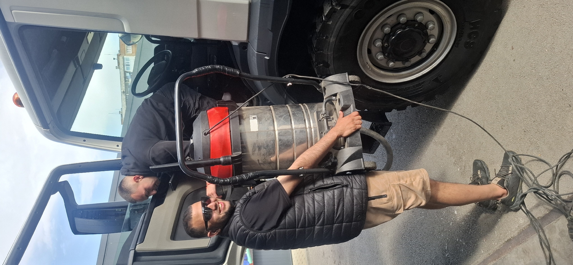 Two workers perform vehicle maintenance outdoors in daylight