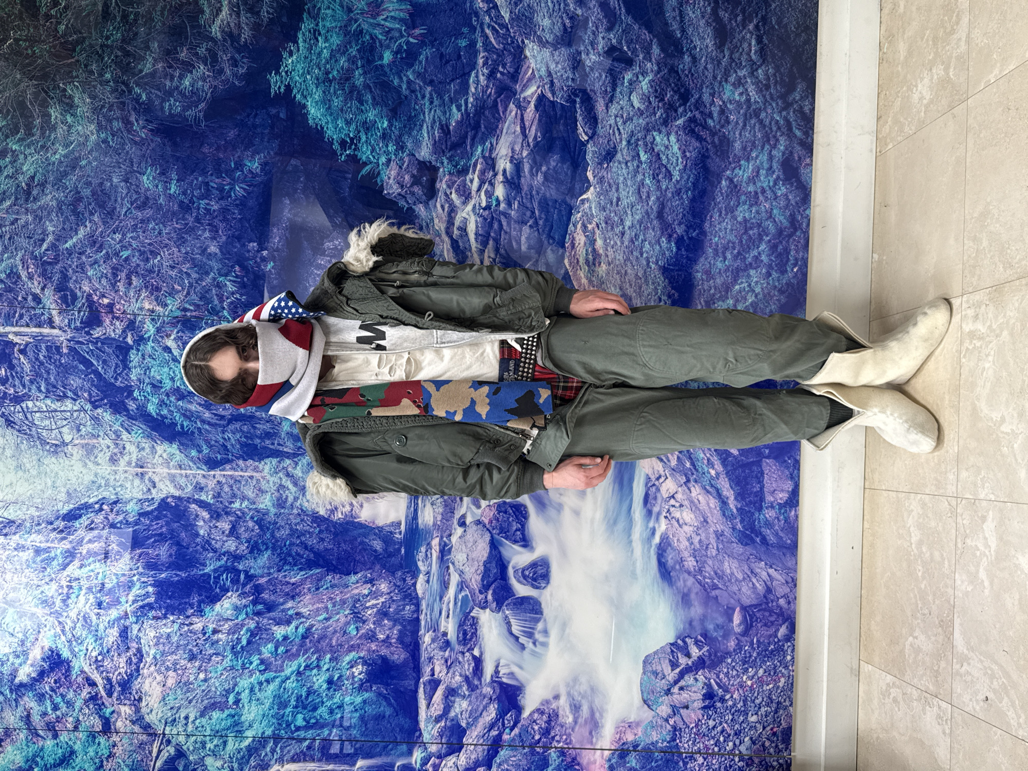 Person photographed in layered clothing against mountain artwork backdrop
