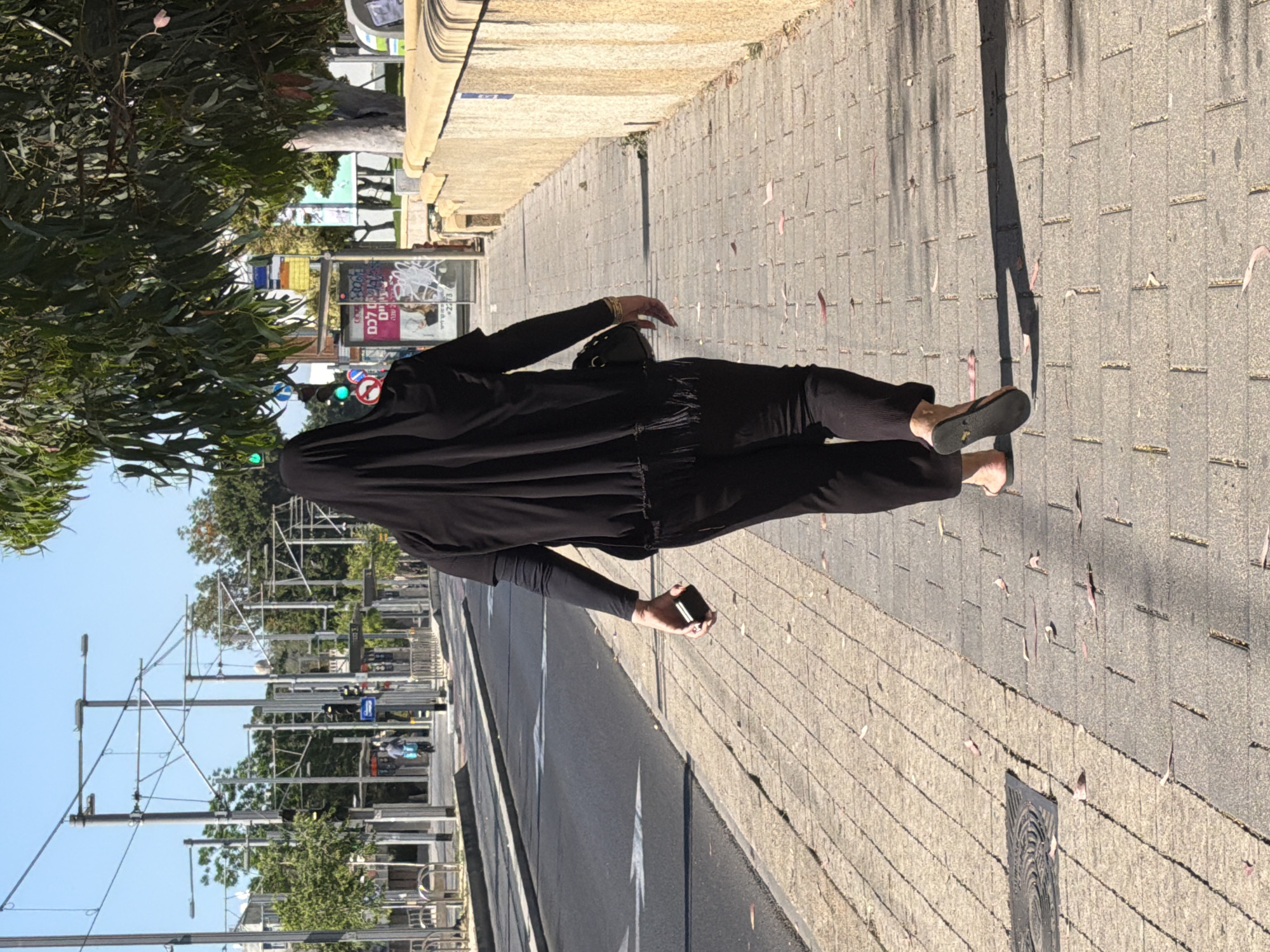 Person in black walks sidewalk with phone in Bat Yam
