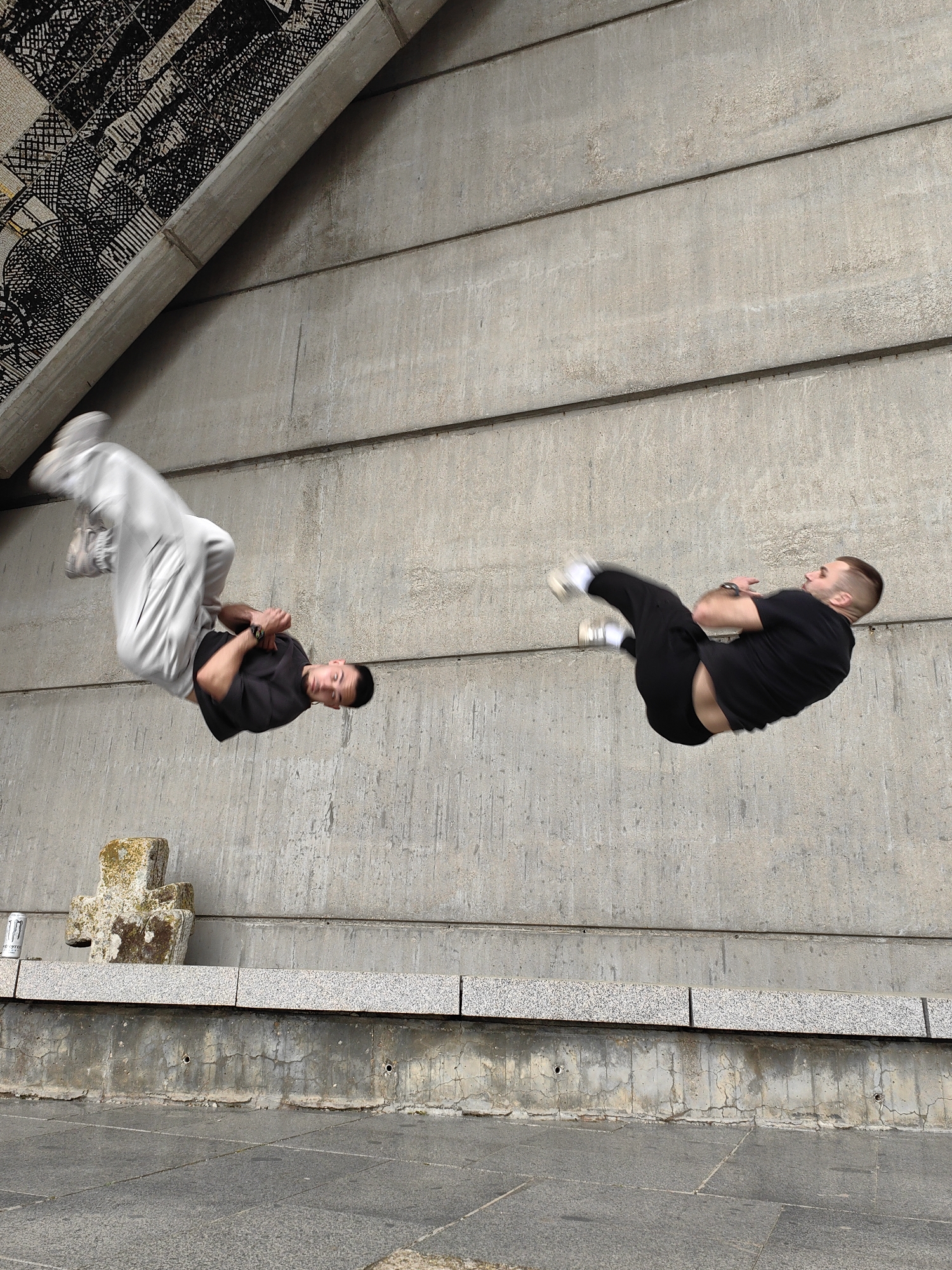 Two men perform synchronized aerial acrobatics near concrete structure