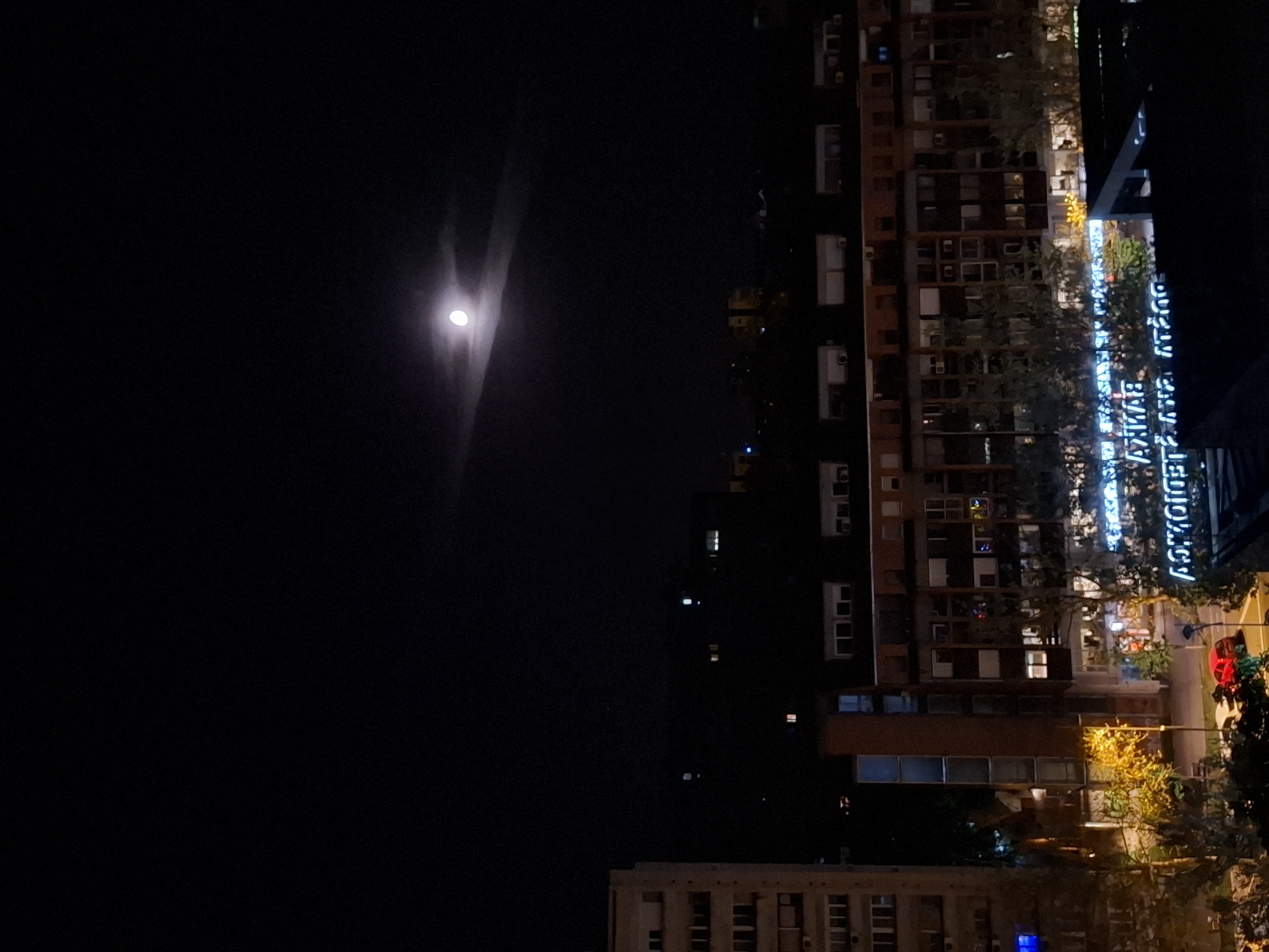 Nighttime moon visible over illuminated apartment buildings in Novi Sad