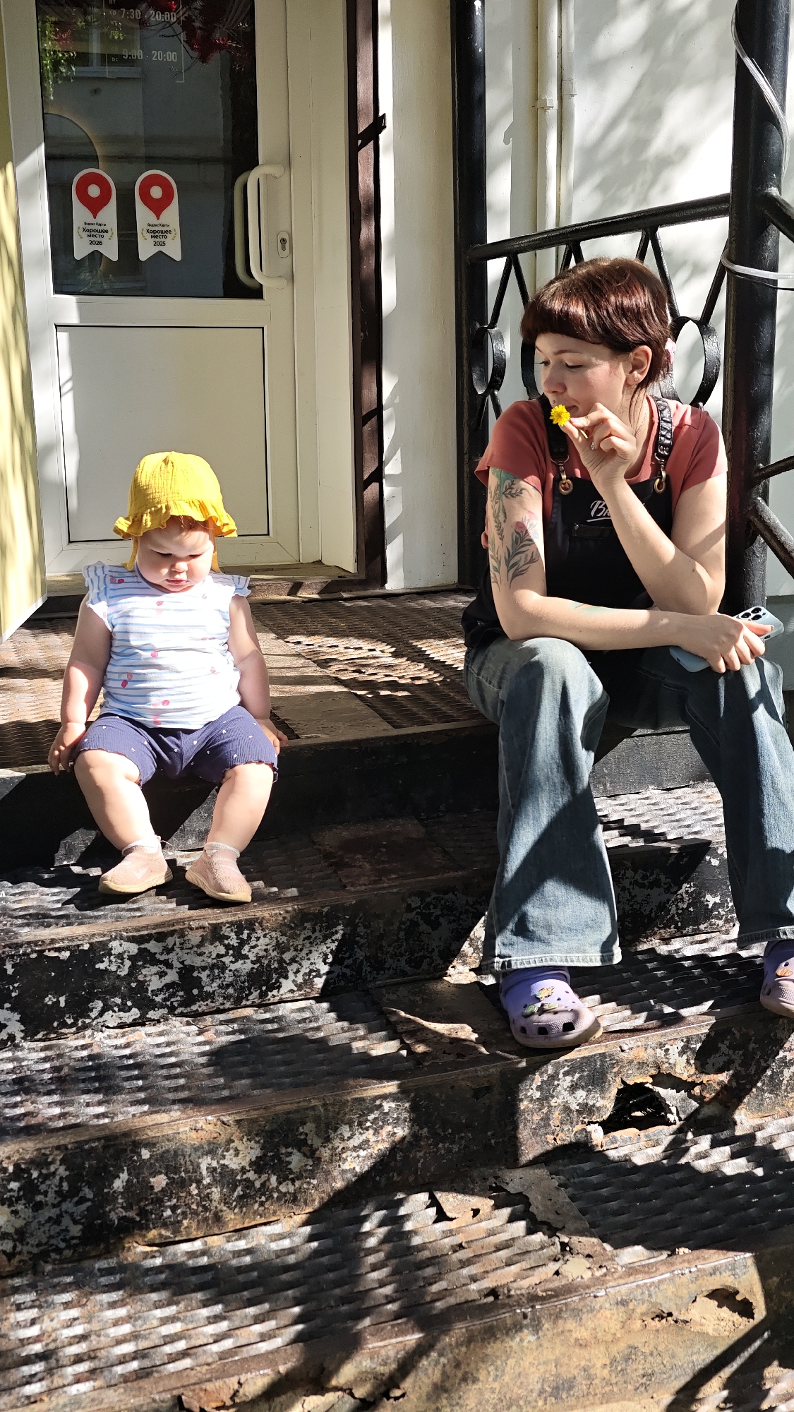 Mother, baby rest outside Chaikovsky business; person holds dandelion nearby