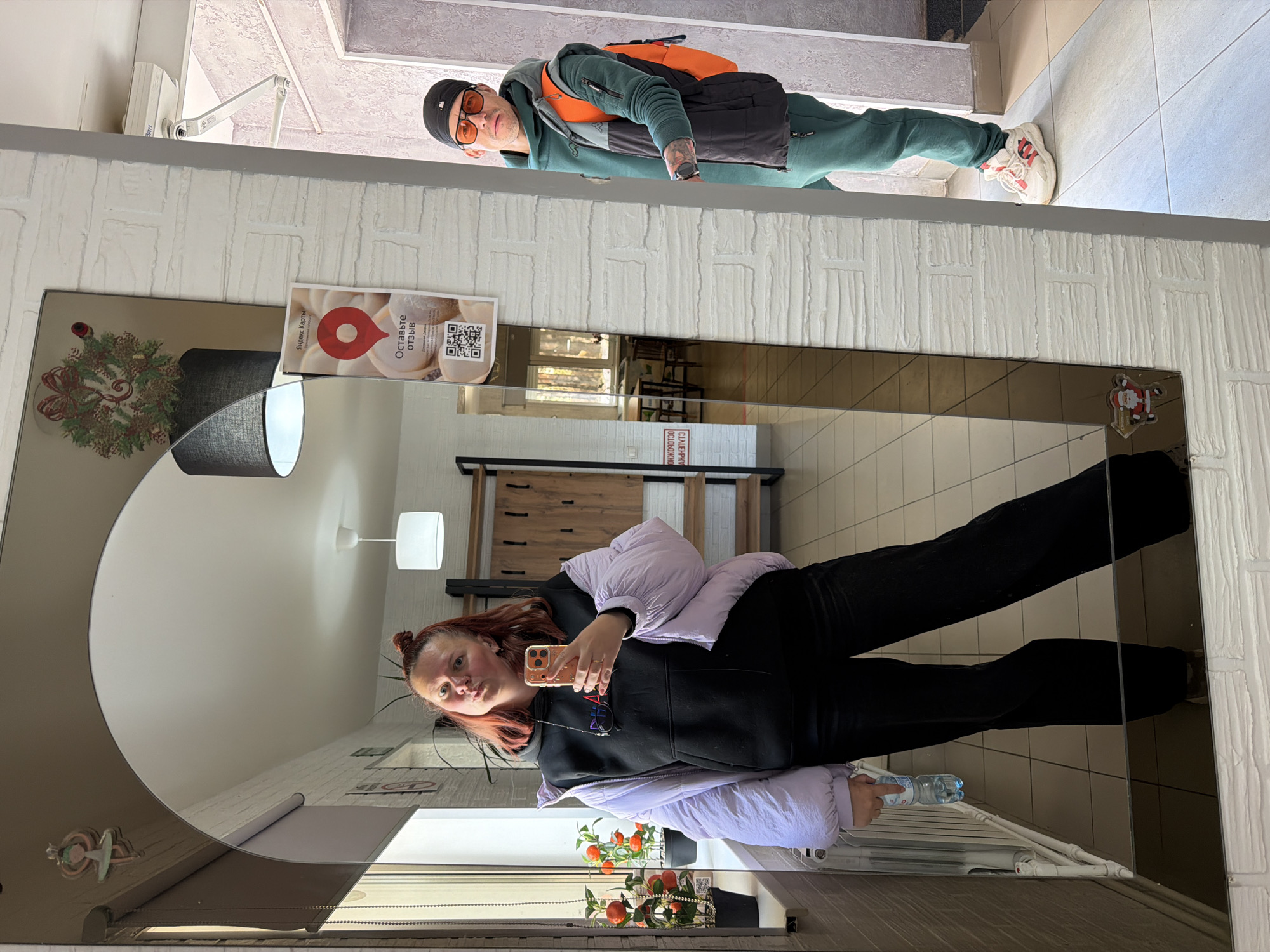 Woman takes mirror selfie with man in background, Noviy