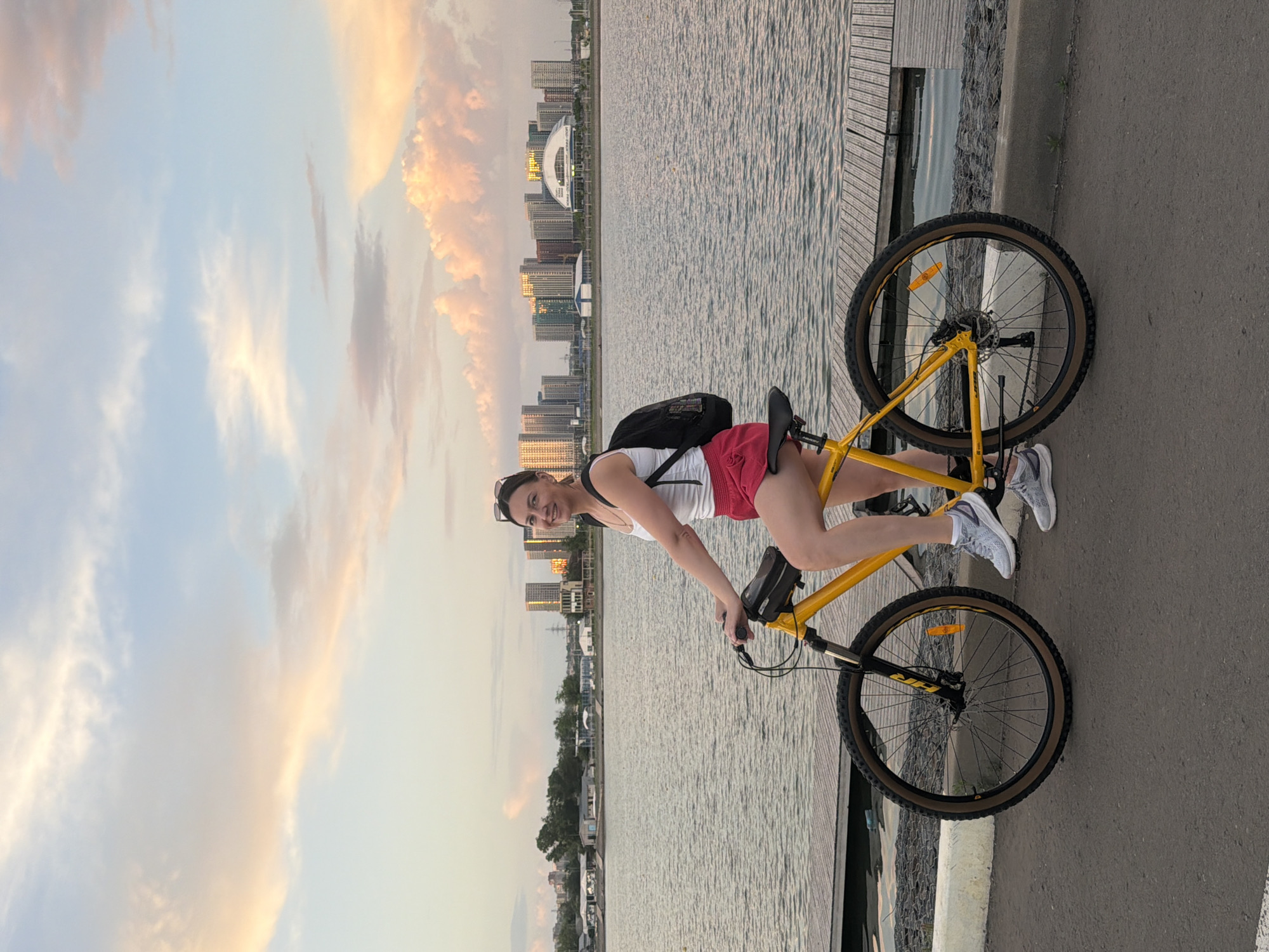Woman poses with bicycle at waterfront during sunset rowing practice