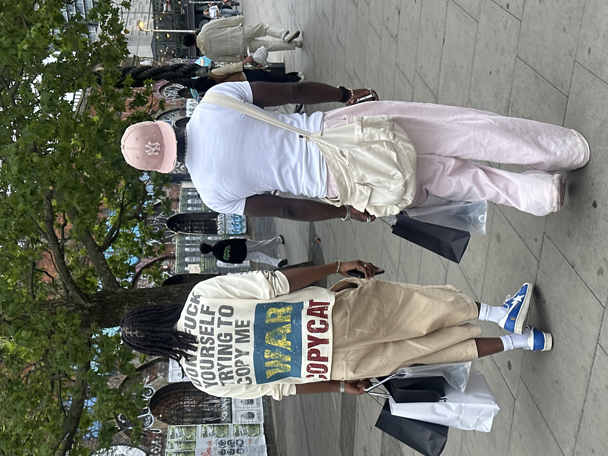 Two pedestrians walk London sidewalk, one wearing slogan shirt