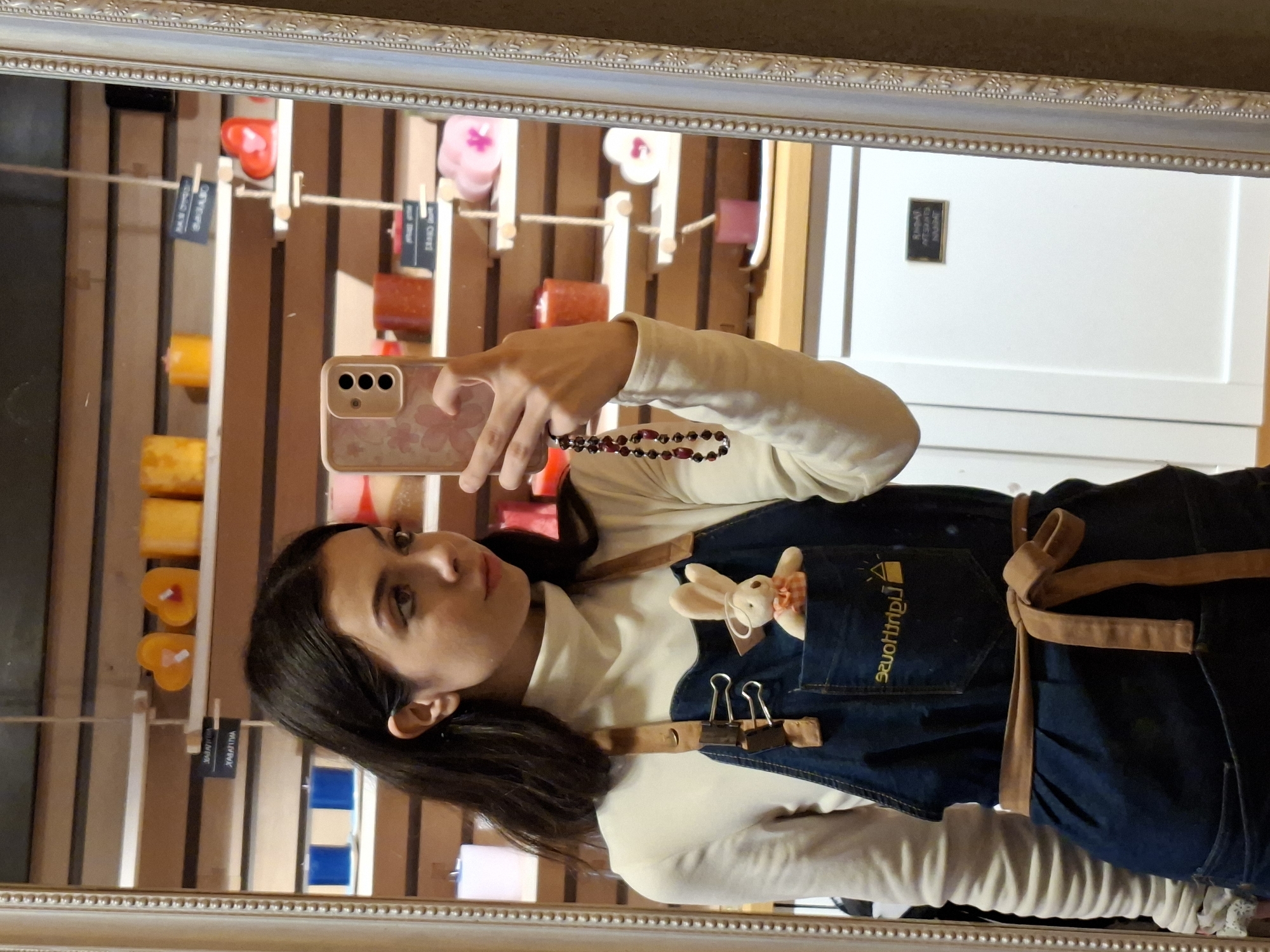 Woman poses in mirror wearing white sweater, denim apron