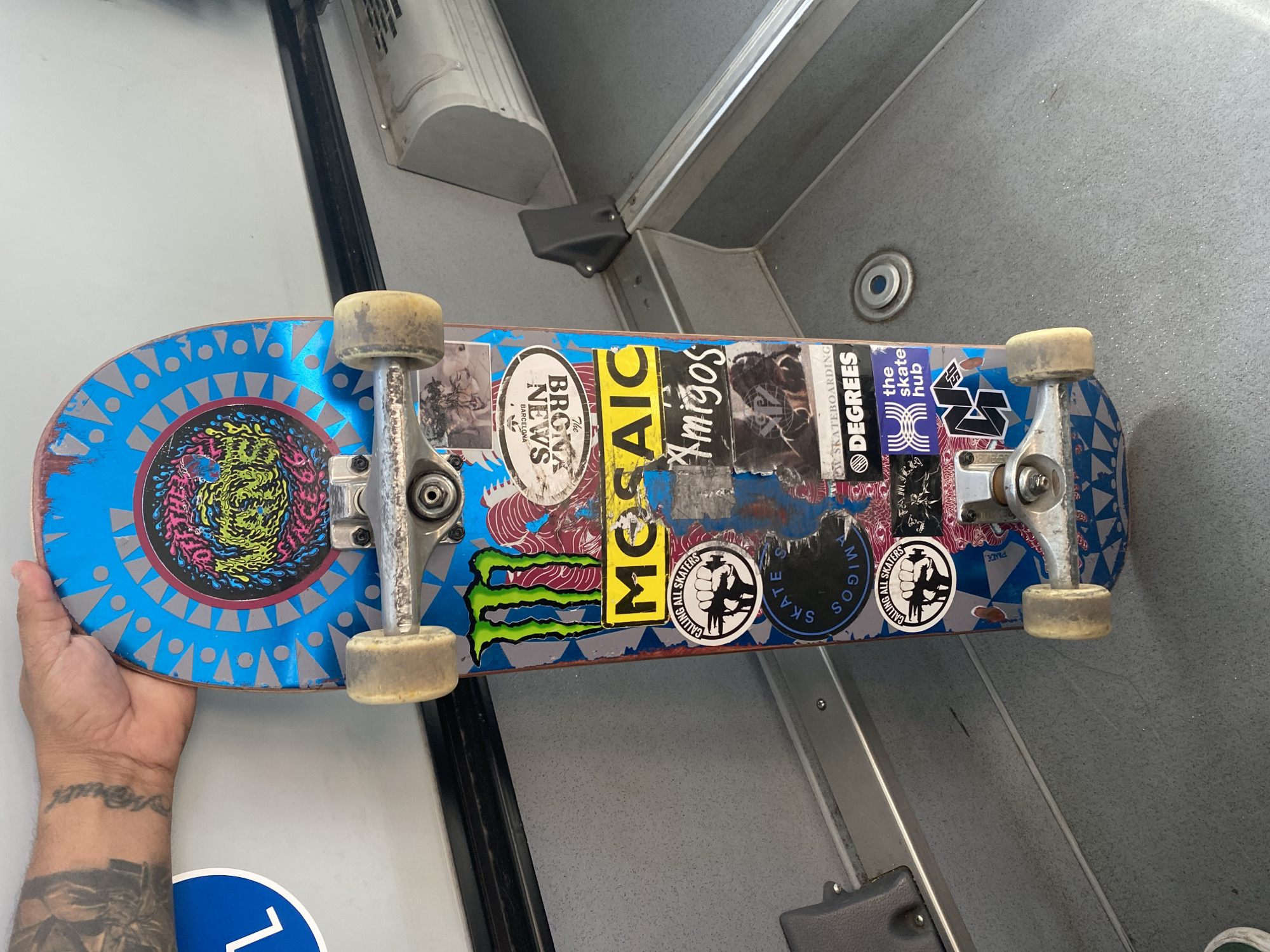 Tattooed skateboarder holds sticker-covered blue deck in Barcelona