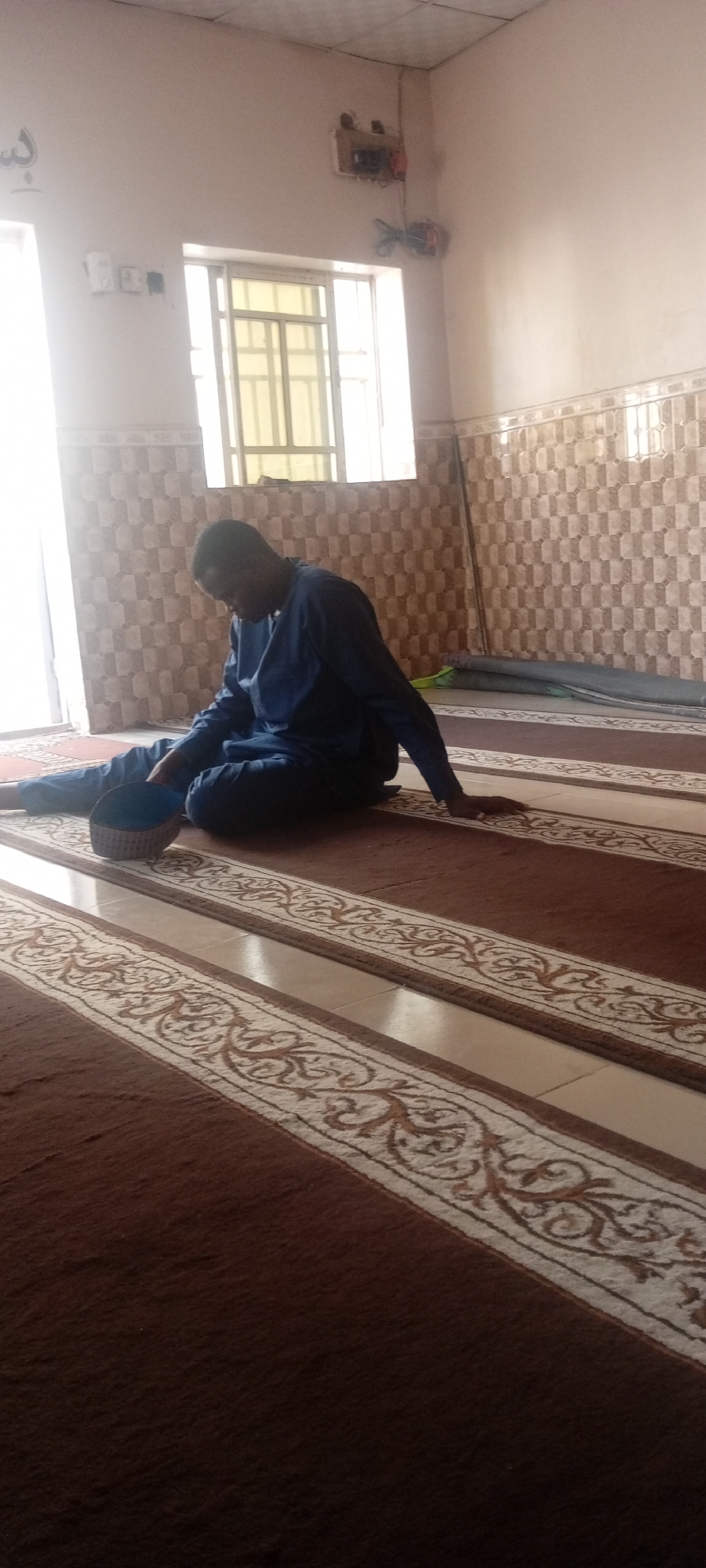 Man in blue tunic sits on patterned carpet in Kano room