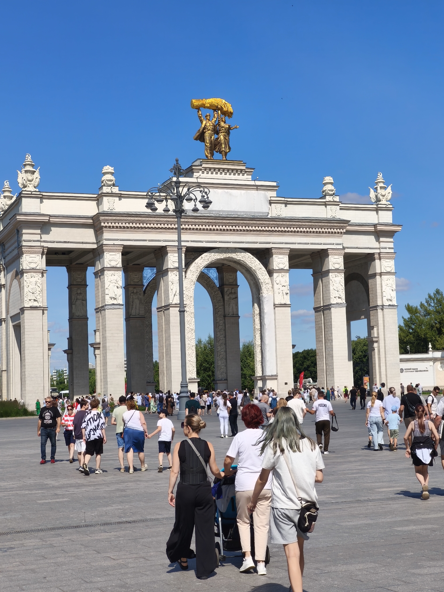 Crowds fill a grand plaza beneath golden wheat sculpture