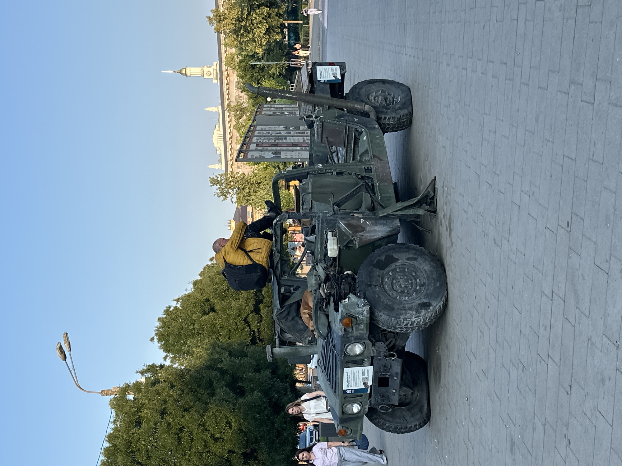 Kyiv street life alongside a displayed war vehicle, summer afternoon