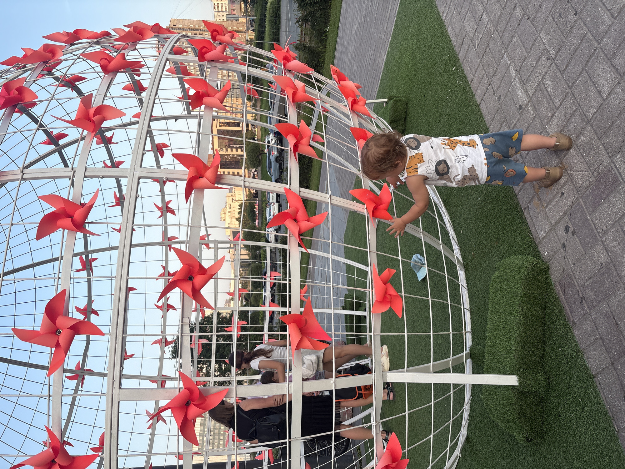 Toddler plays around red pinwheel sphere and water jets in urban plaza