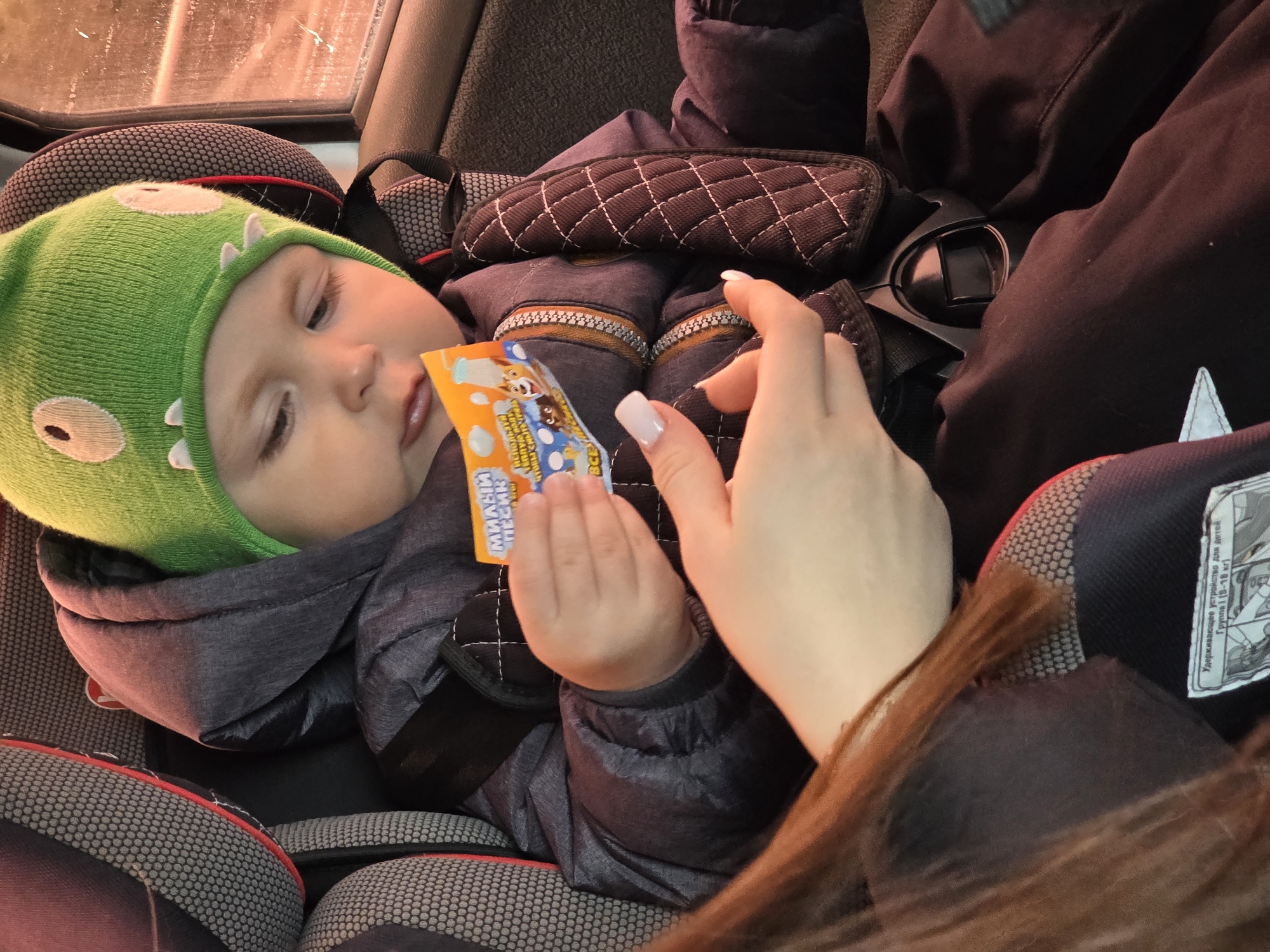 Baby in cartoon hat holds packet during car ride