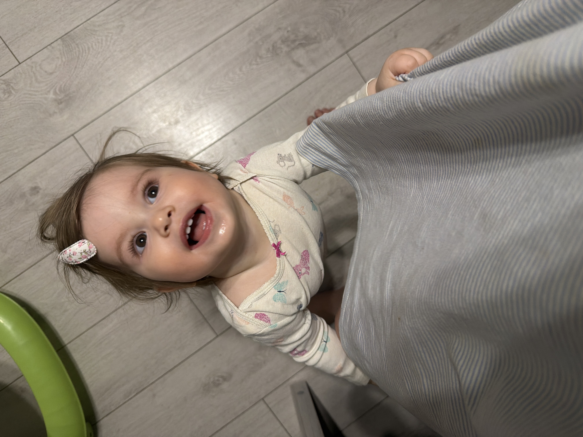 Child in onesie holds curtain on wooden floor
