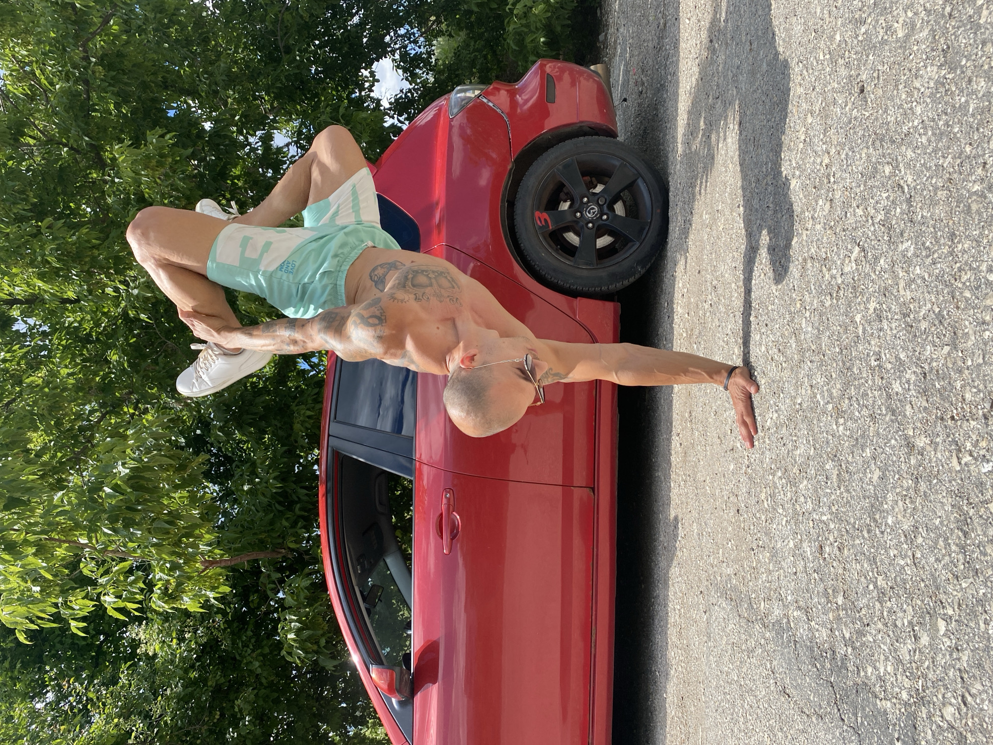 Tattooed figure poses with baseball bat near red car in Pleven