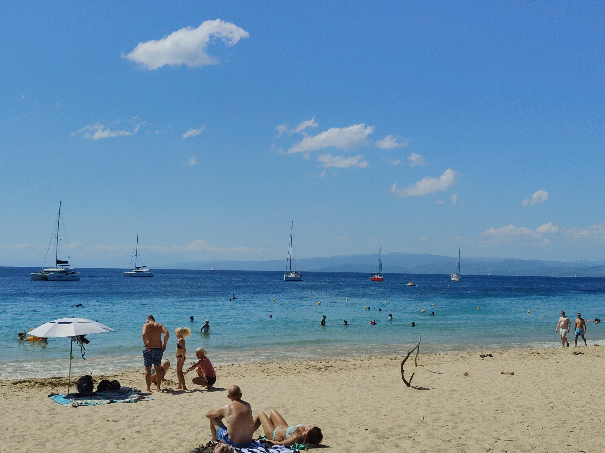 Busy Greek beach morning — swimmers, cafe, sailboats, coastal flora