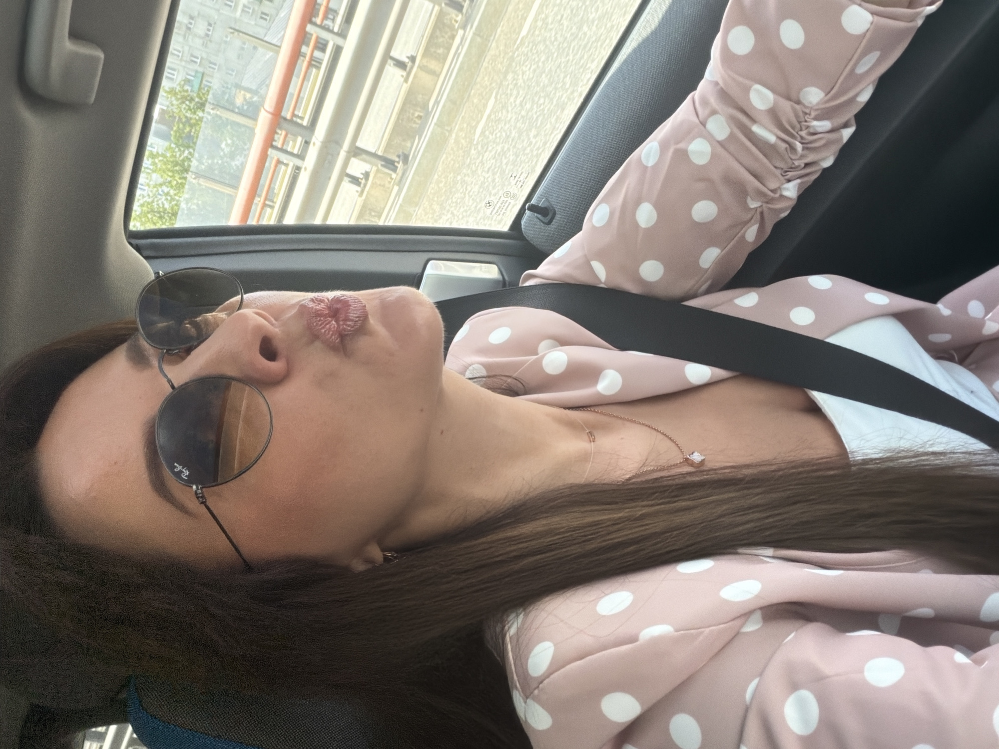 Inside a car in Riga, pink blazer, city streets rolling by.