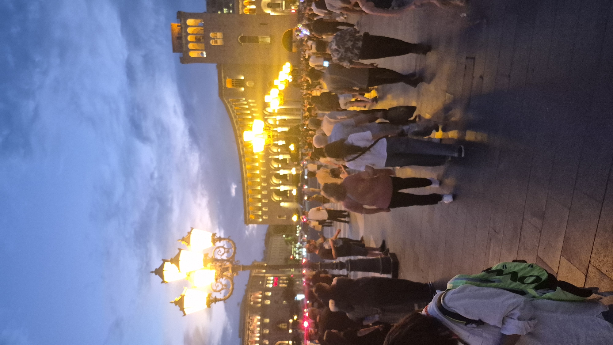 City square at dusk, crowds moving under glowing streetlamps