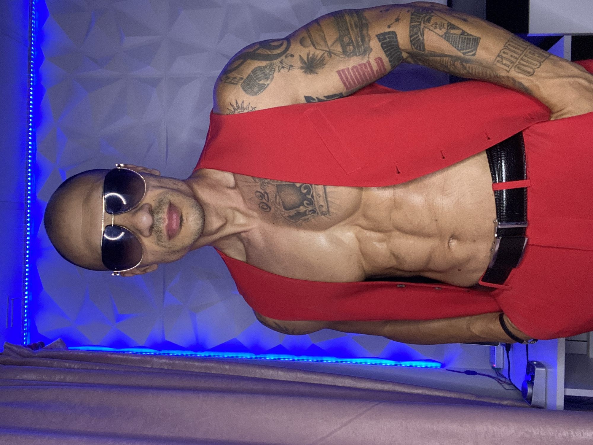 Tattooed figure in red stands against a textured wall in Pleven
