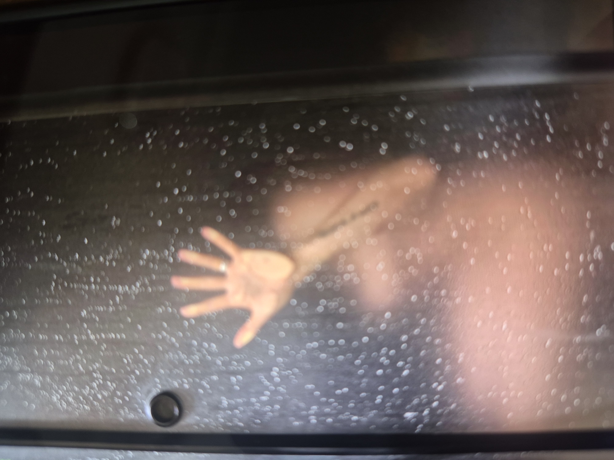 Figure observed behind wet glass surface, hand pressed against barrier