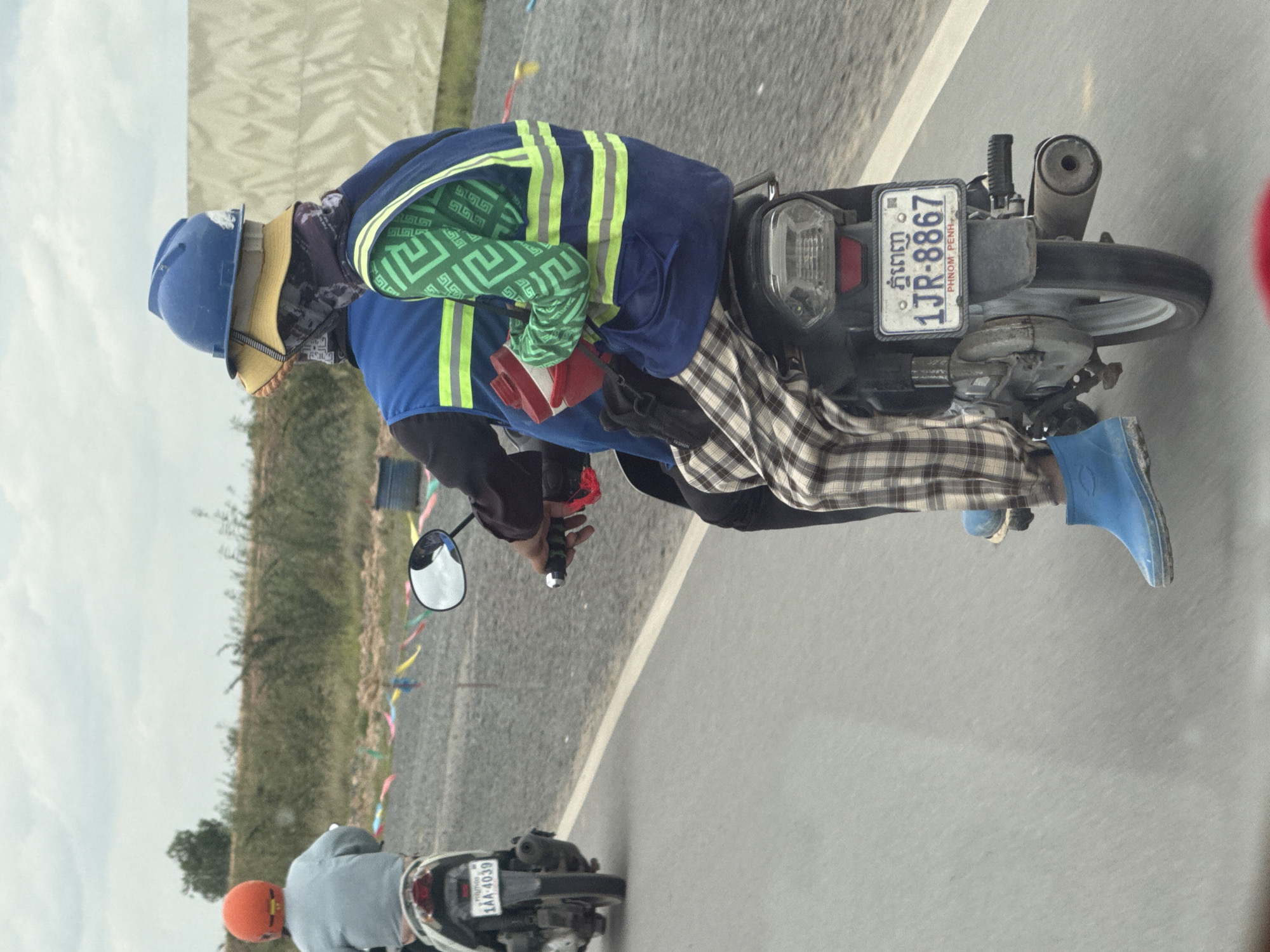 Motorcycle riders navigate Krong Ta Khmau streets, layered up against the elements