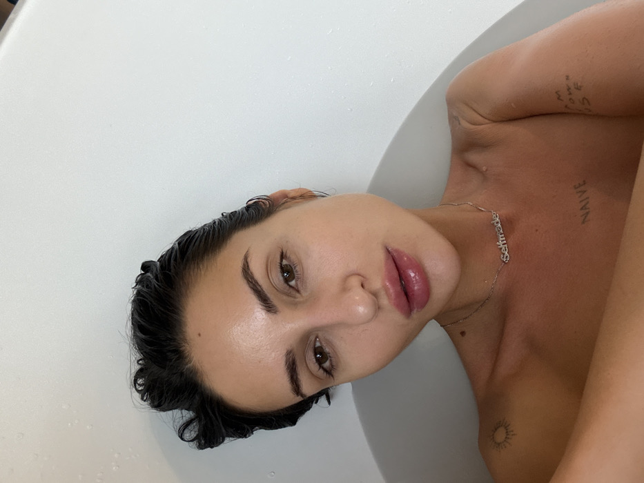 Woman observed in bathtub wearing silver necklace, tattoos visible