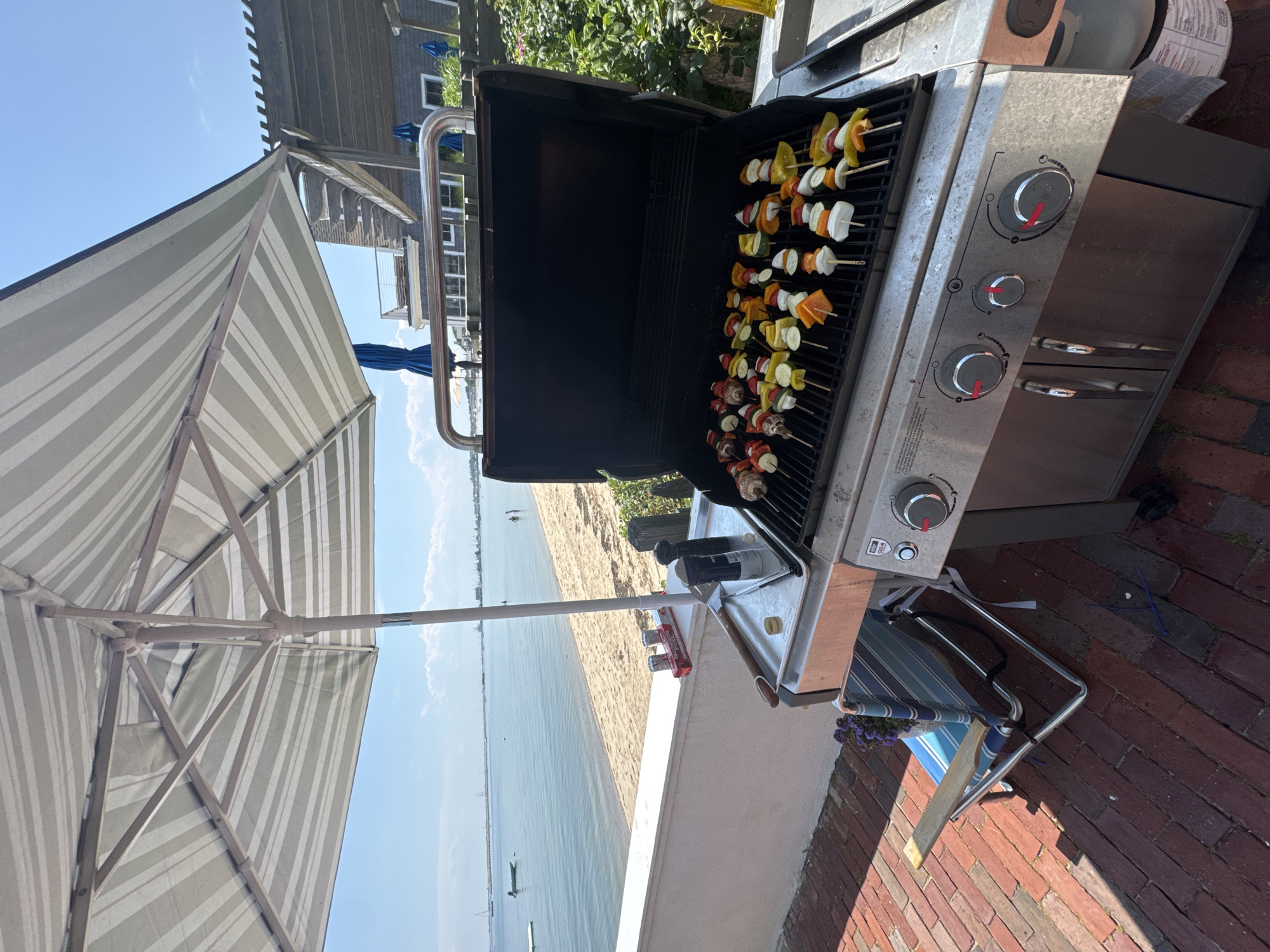 Vegetable skewers on an open barbecue grill under a striped umbrella