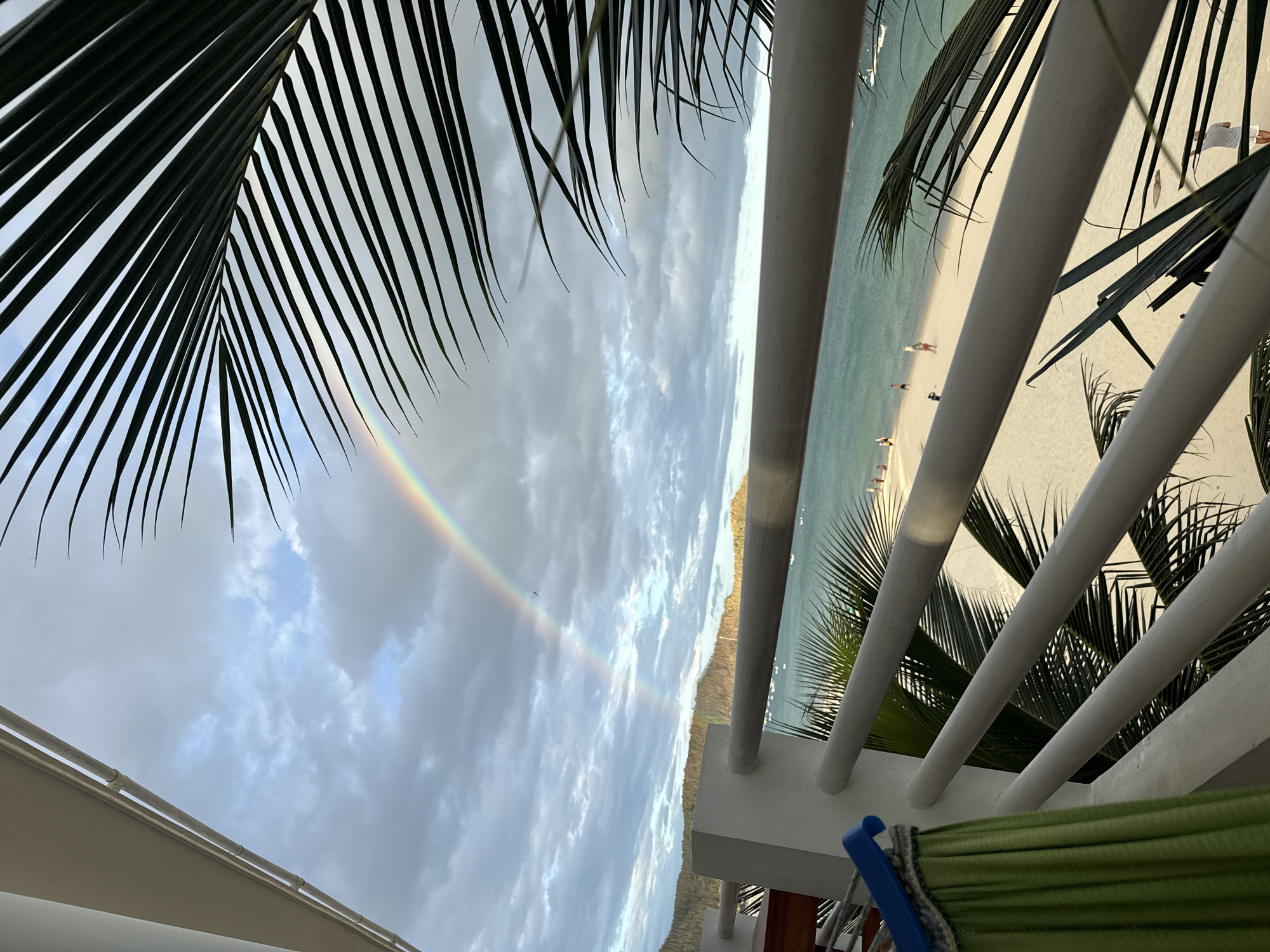 Double rainbow over a tropical beach, seen from a palm-framed balcony