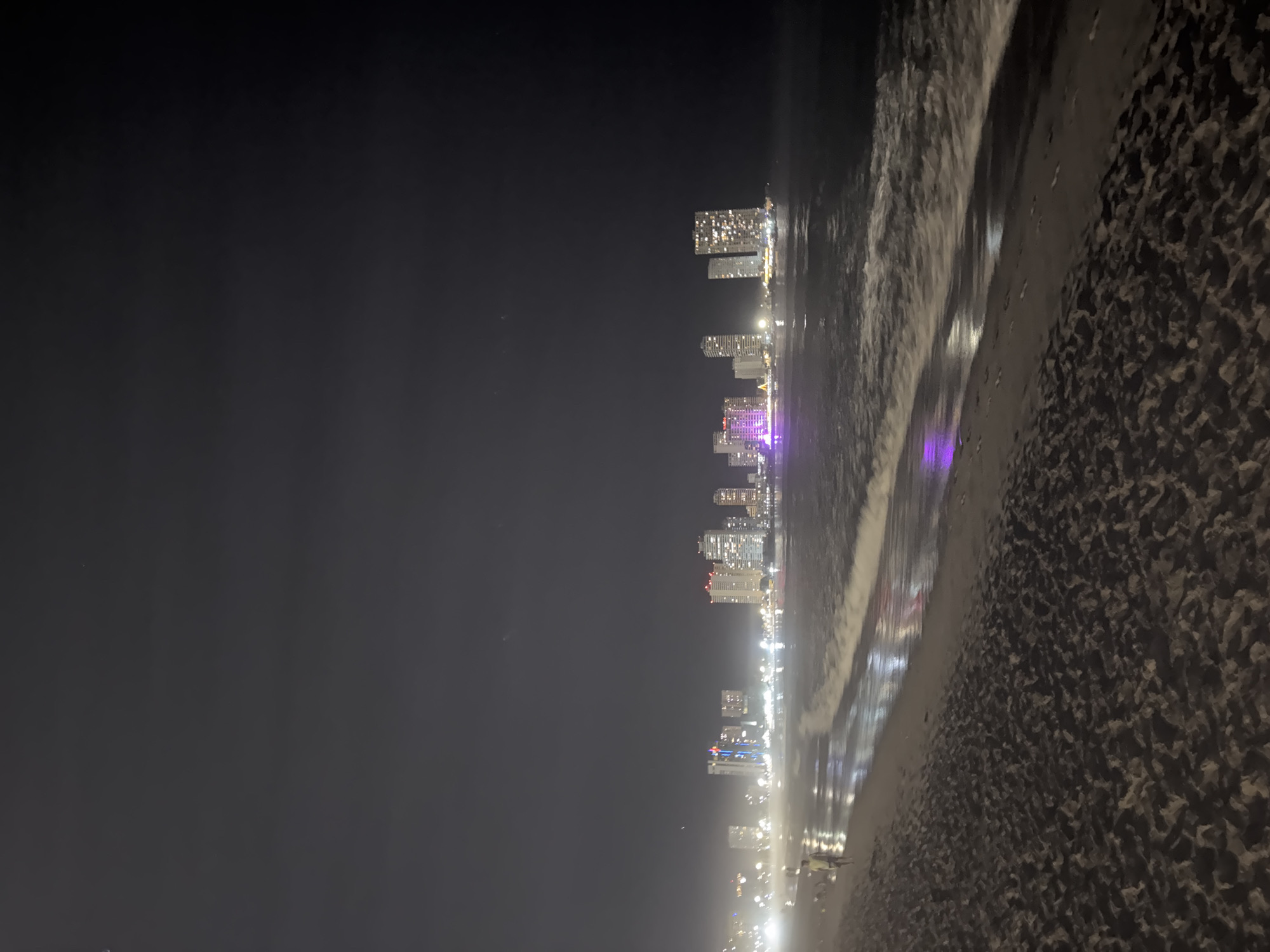 Nighttime beach scene with distant city skyline, lone figure observed