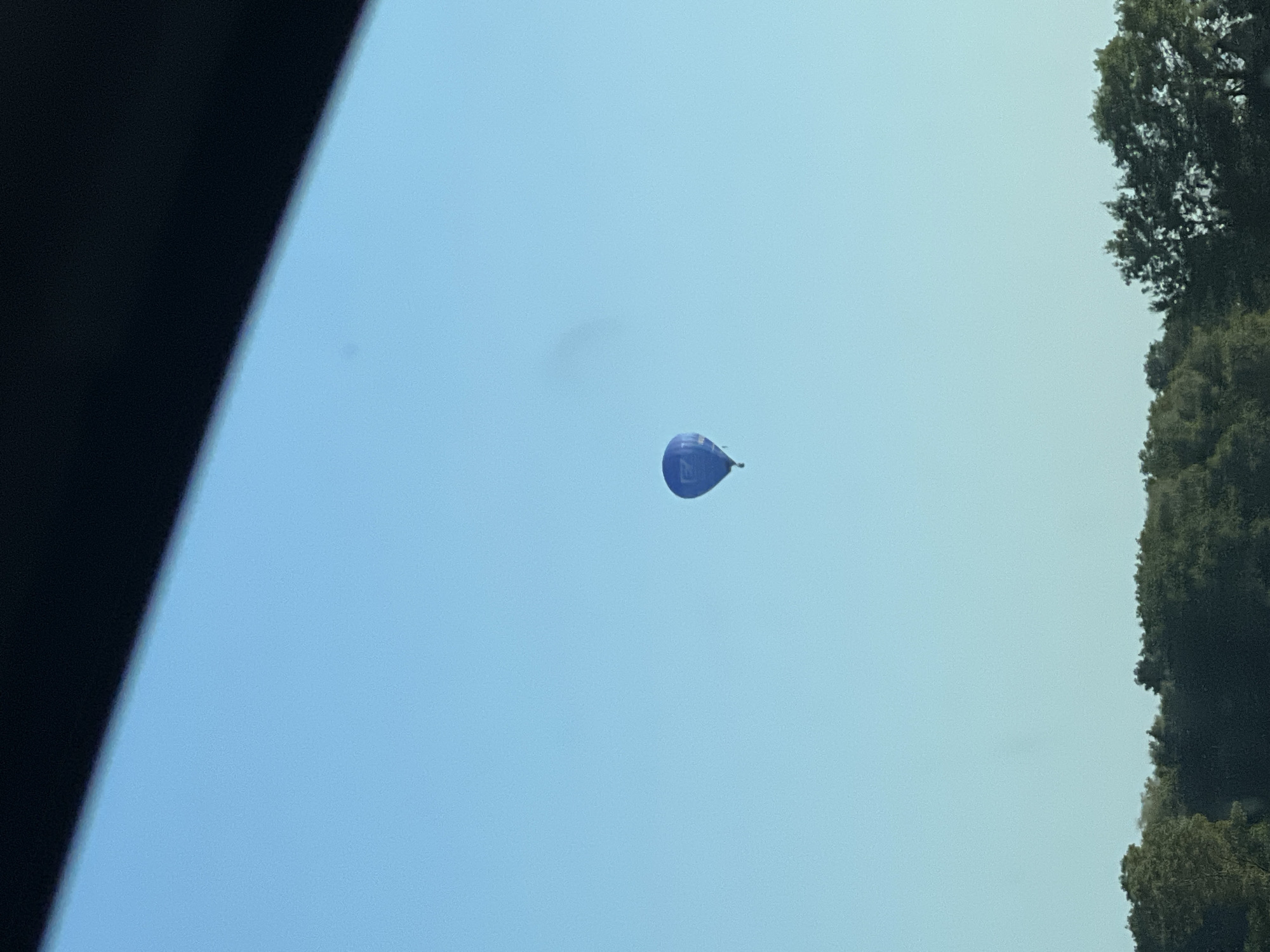 Hot air balloons drift over treetops as a logging truck waits at a red light