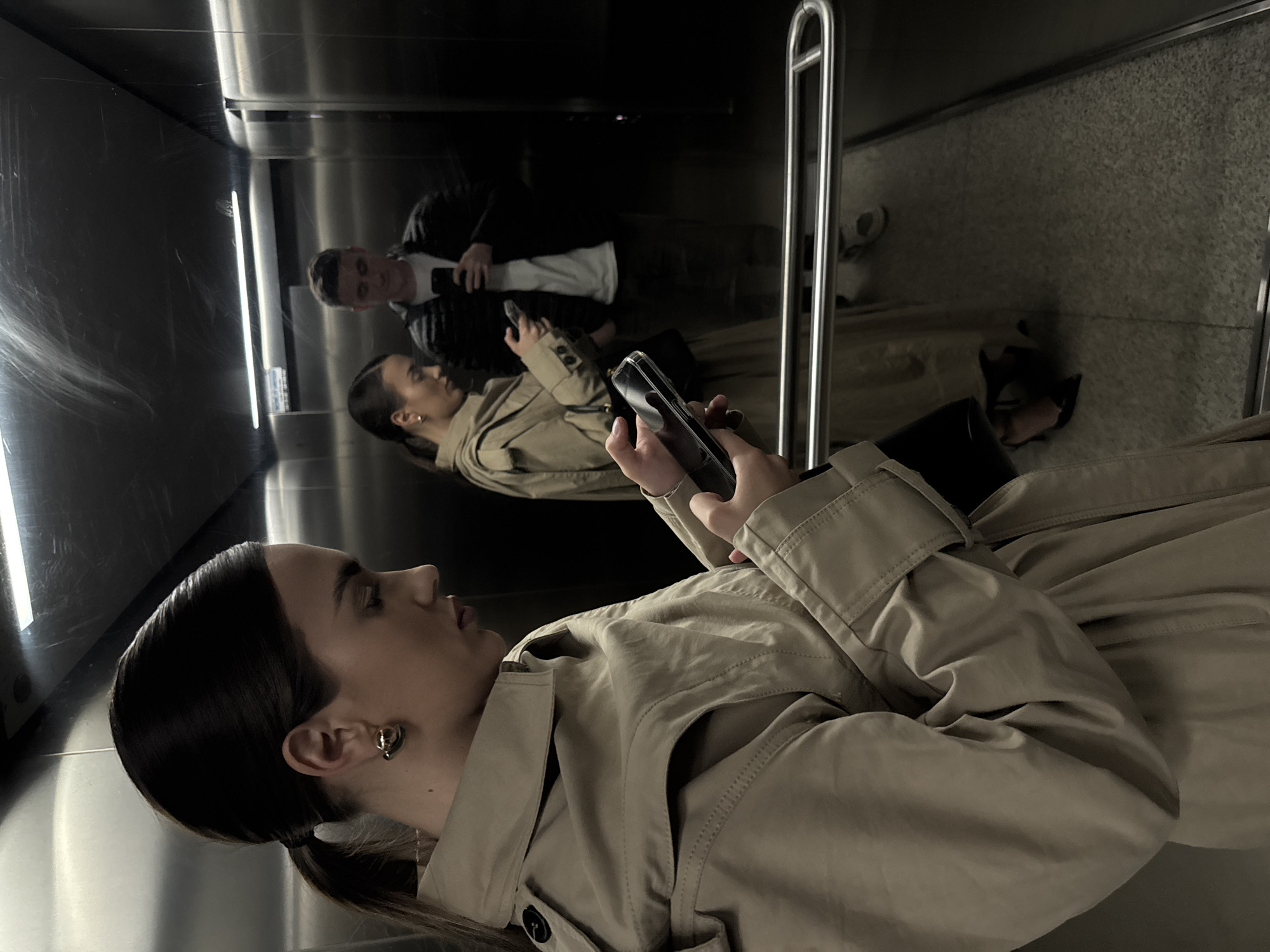 Woman in trench coat uses phone in building hallway