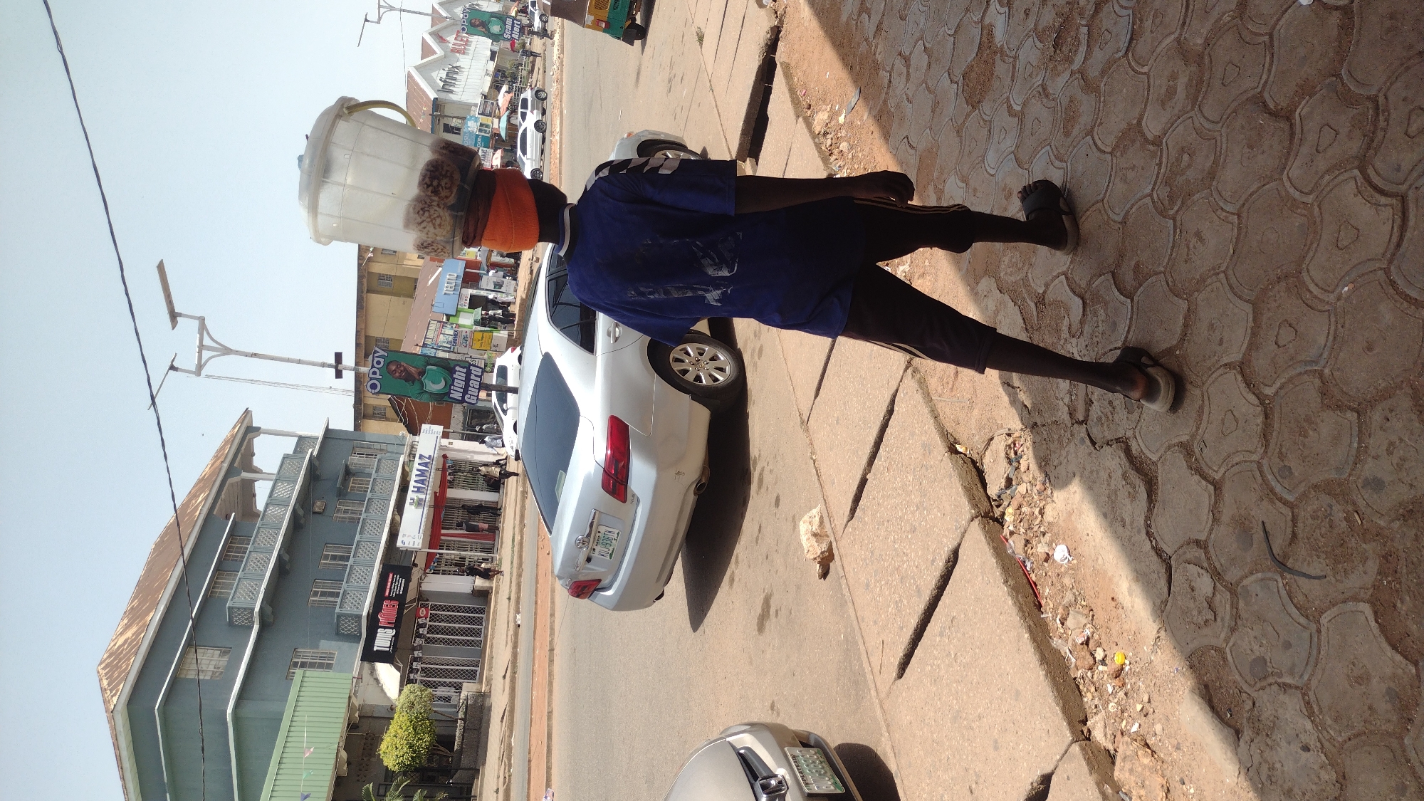 Person carries container on head along Jos sidewalk