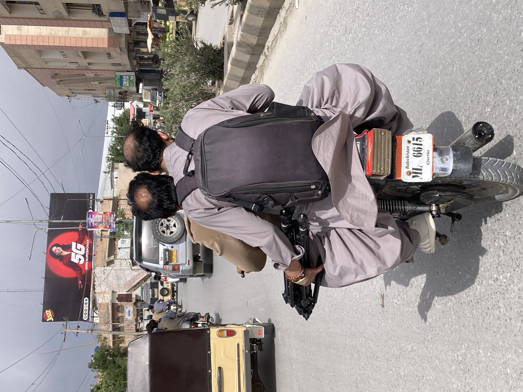 Two men ride motorcycle through Quetta streets under overcast sky