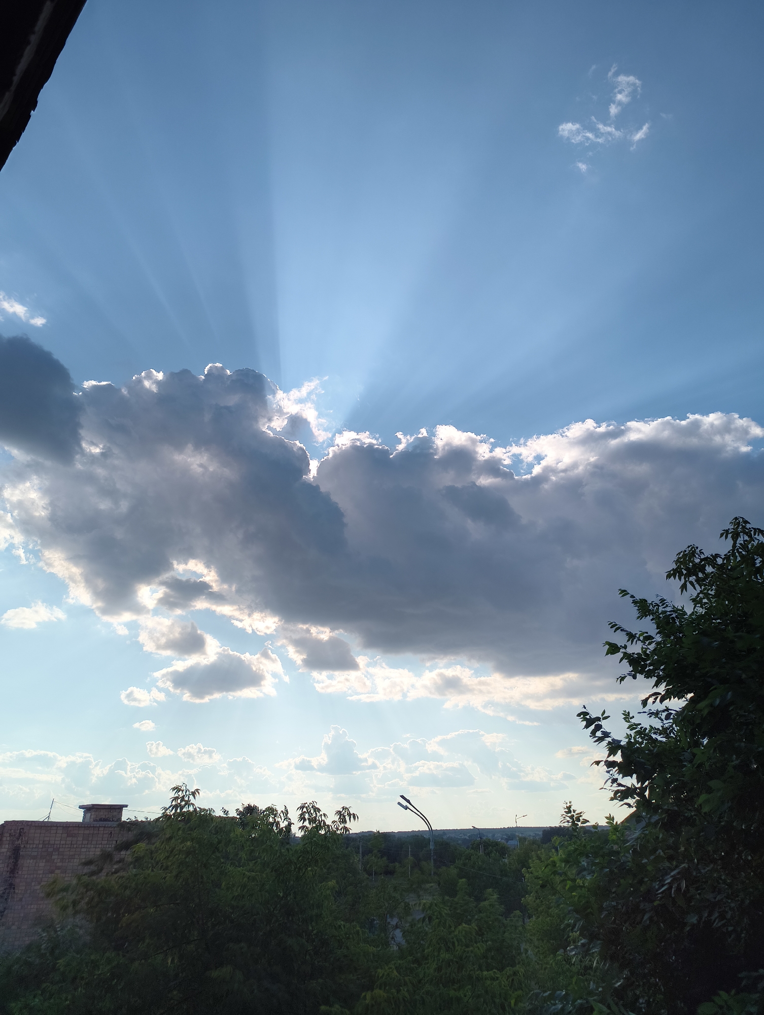 Crepuscular rays break through clouds above a tree-lined cityscape