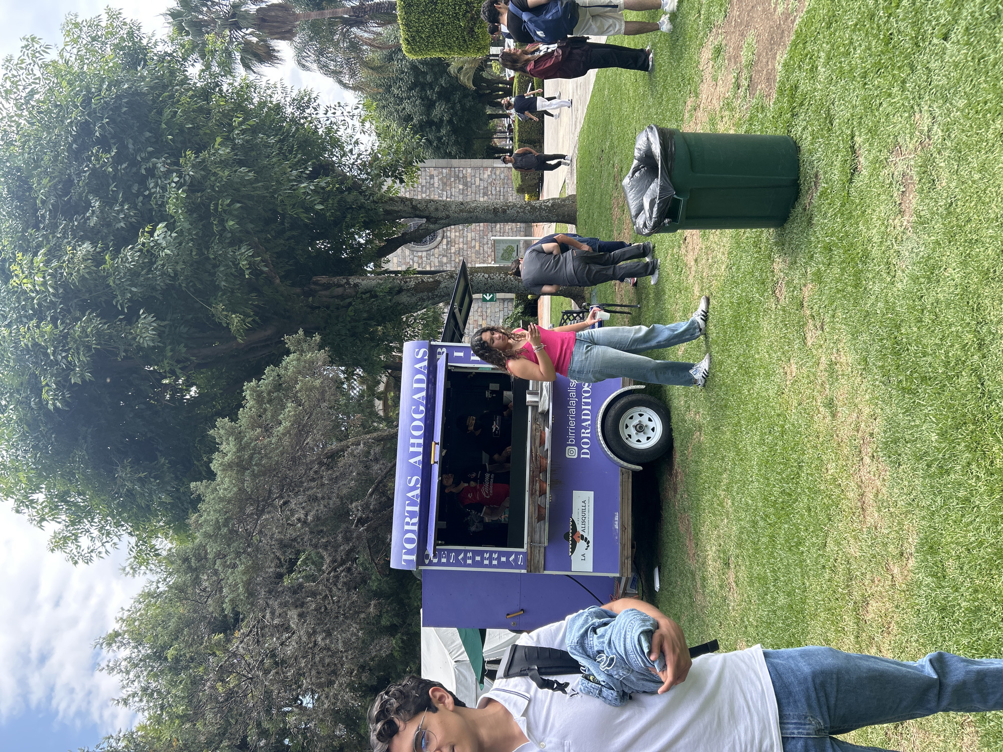 Food vendors serve customers amid blooming gardens and Jacaranda trees