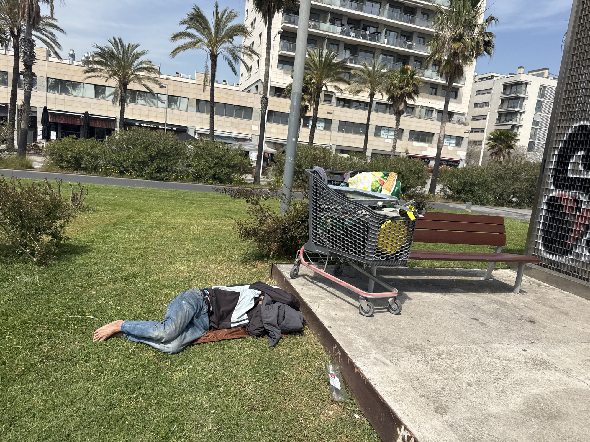 Children skate while homeless man rests at Barcelona waterfront park