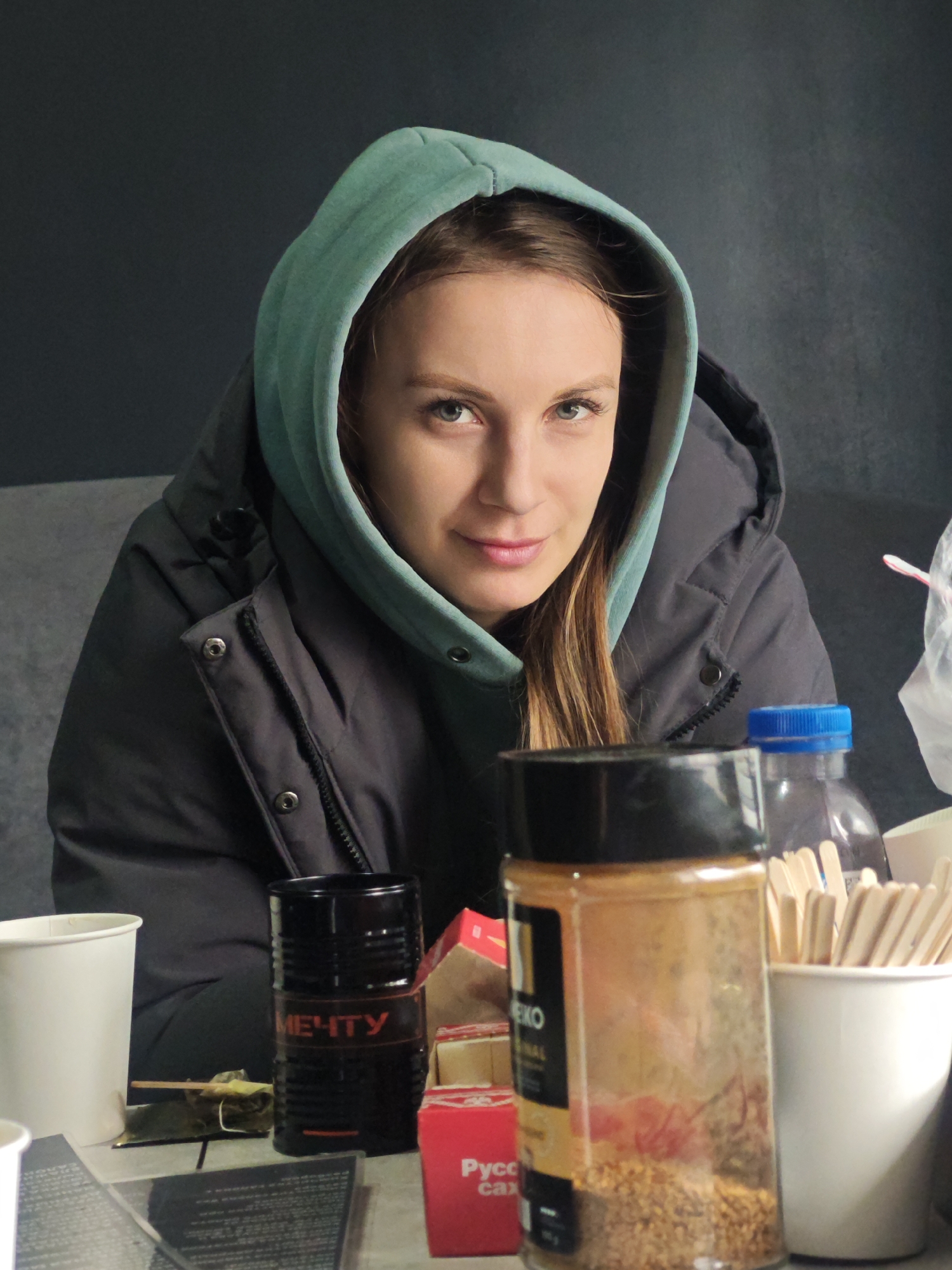 Young woman in green hoodie sits at cluttered table