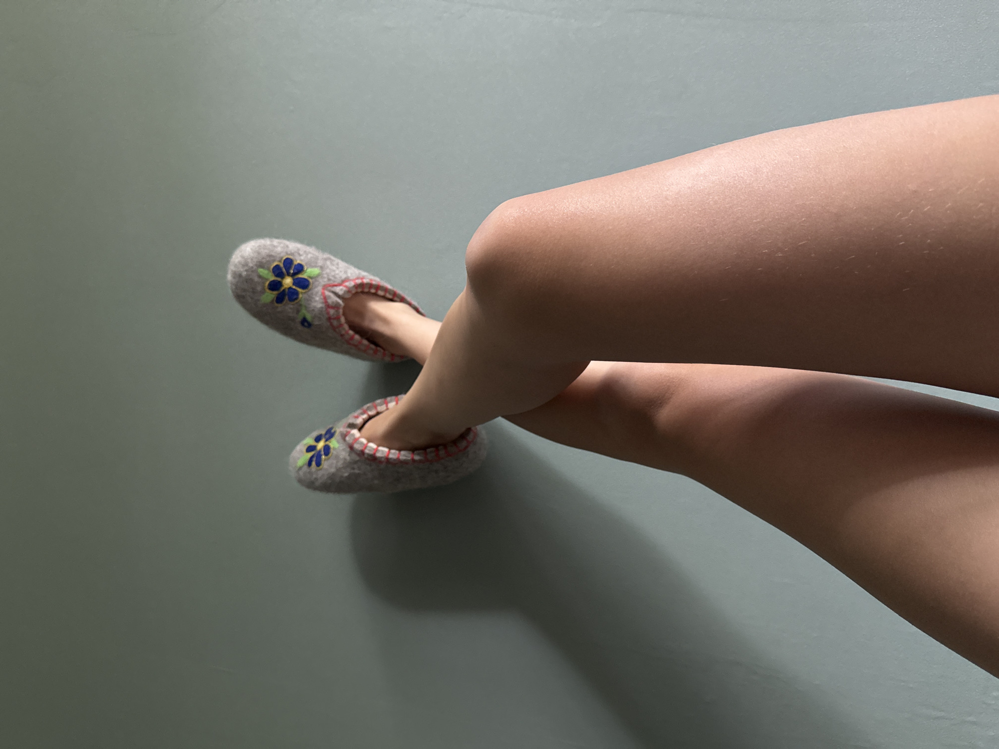 Relaxing indoors in embroidered felt slippers against a teal wall.