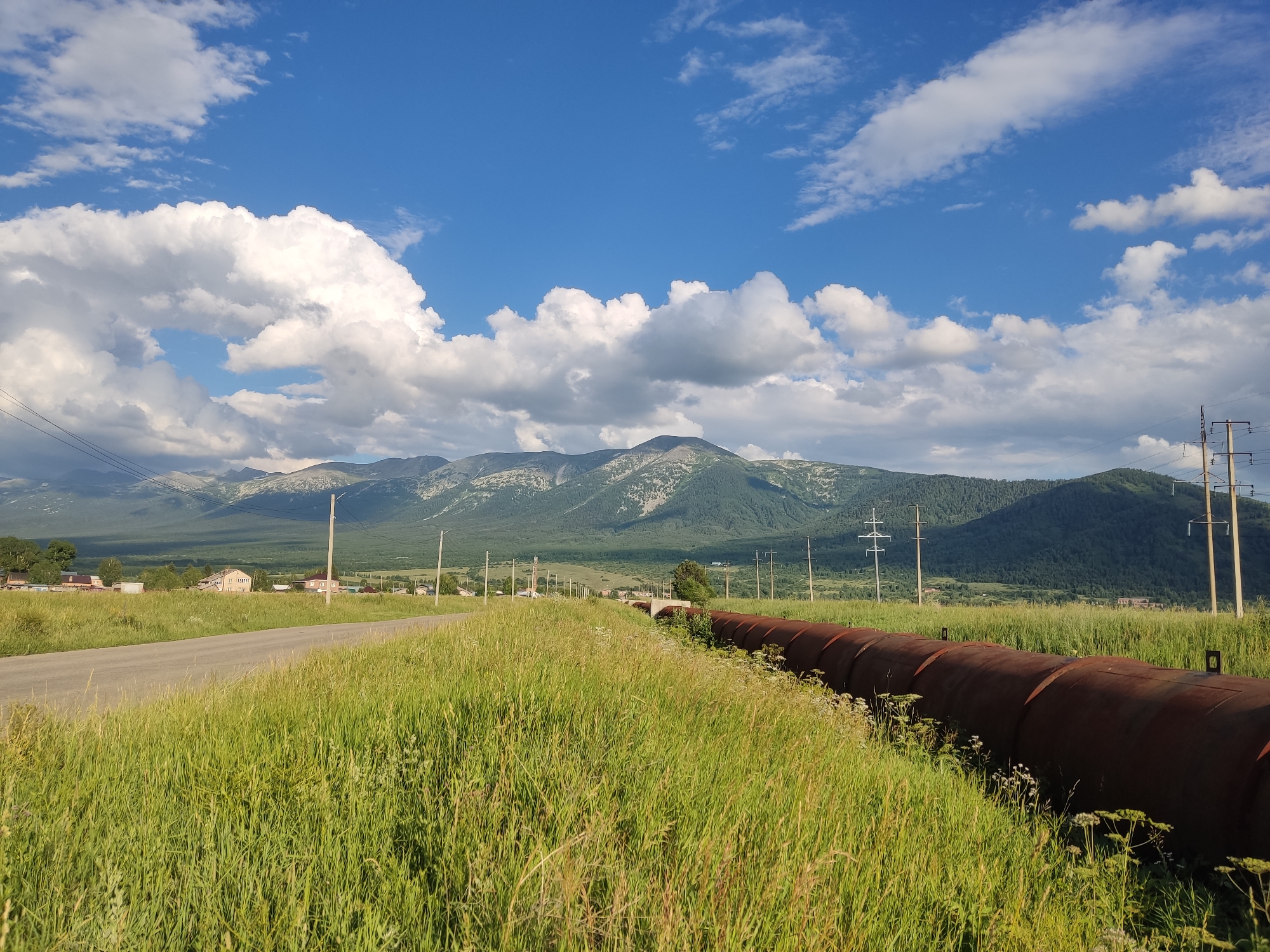 Rural road, rusty pipeline, and Soviet-era war memorials in open countryside