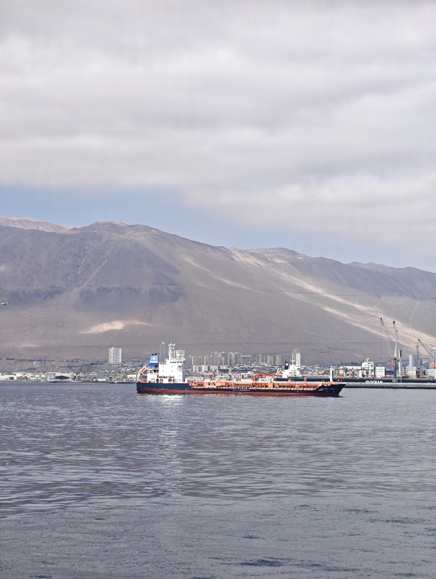 Cargo ship passes a mountainous coastline under heavy overcast skies.