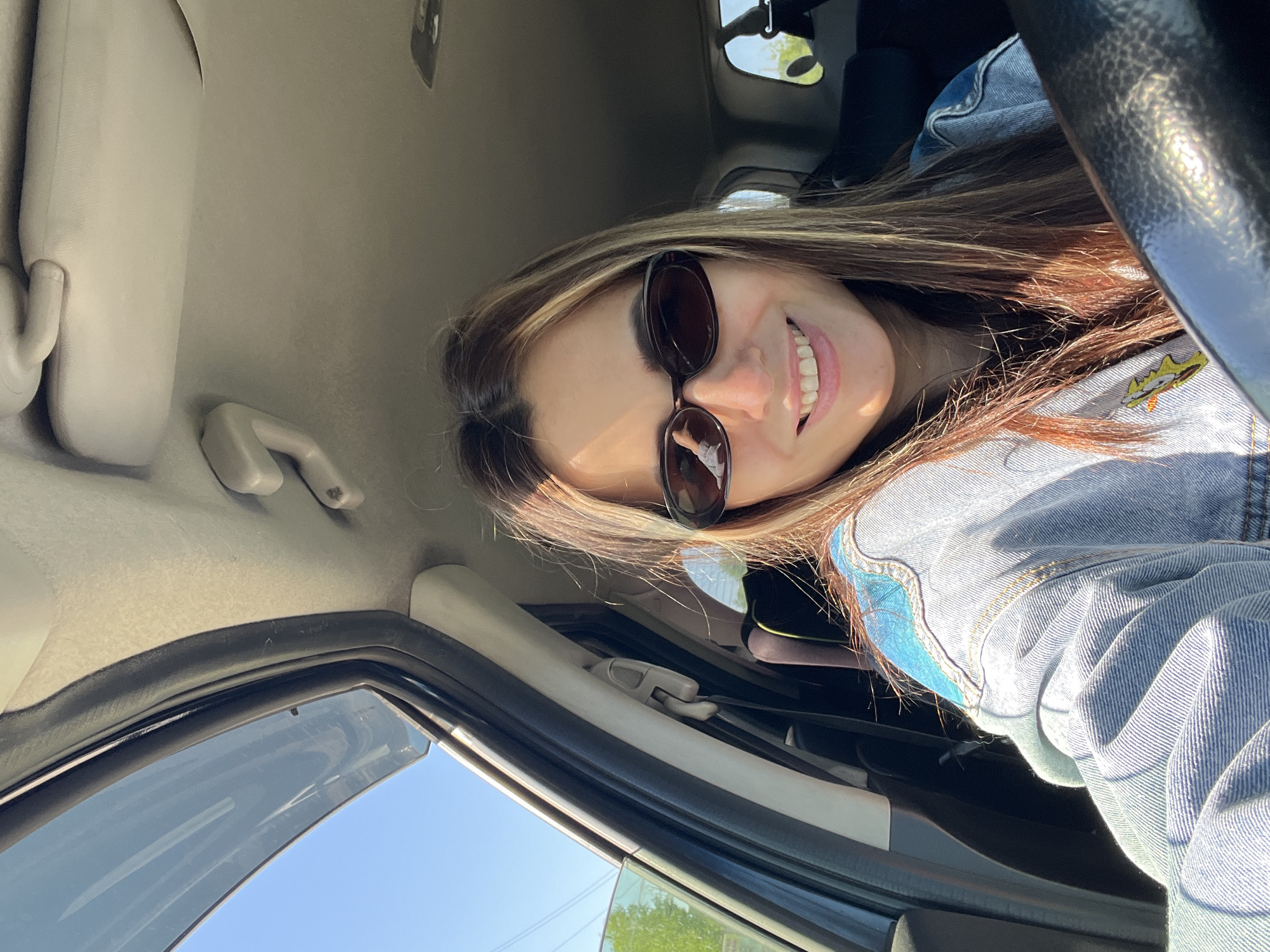 Woman photographed in car wearing denim jacket and sunglasses