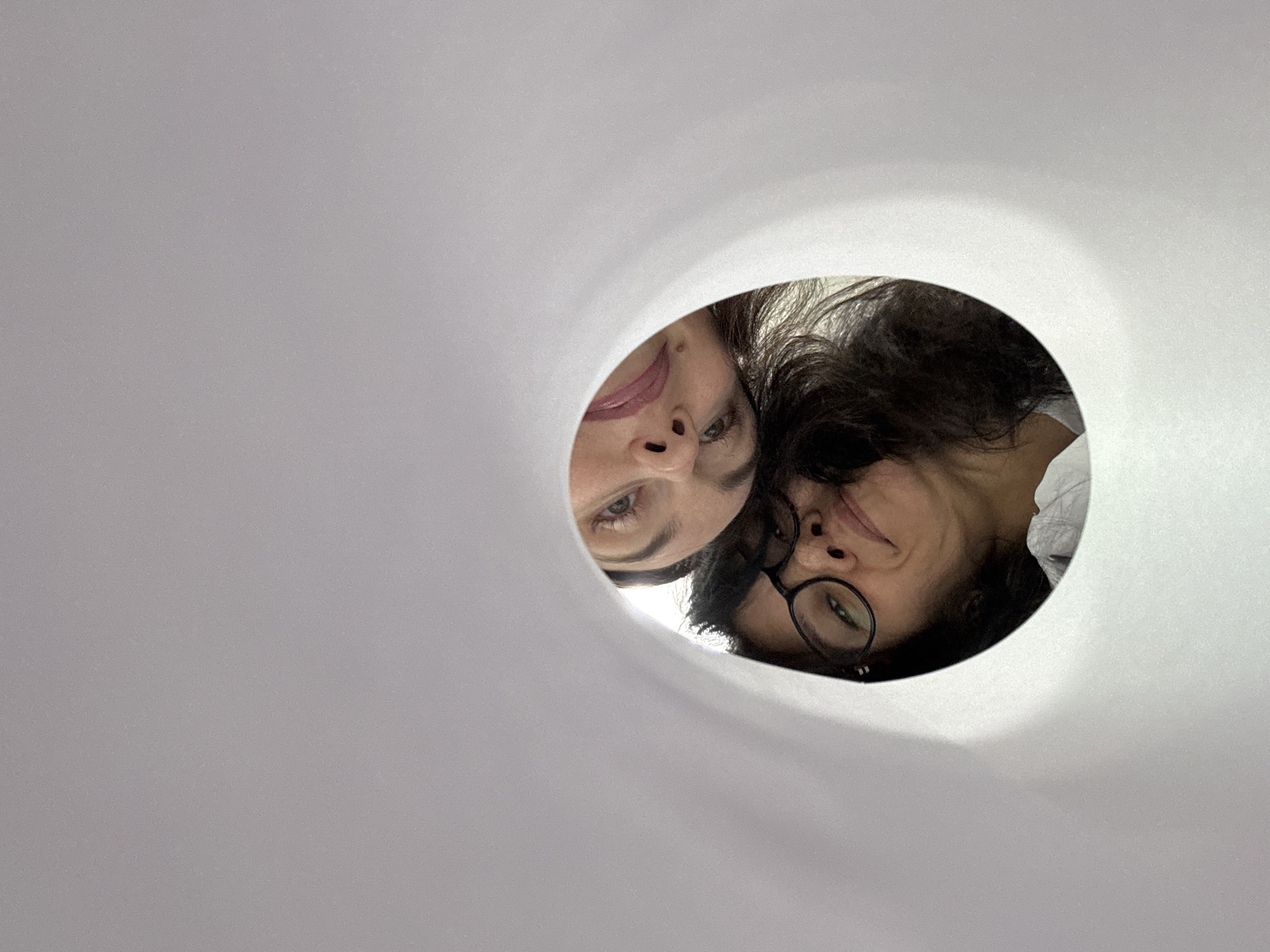 Two people peer through circular opening in white surface