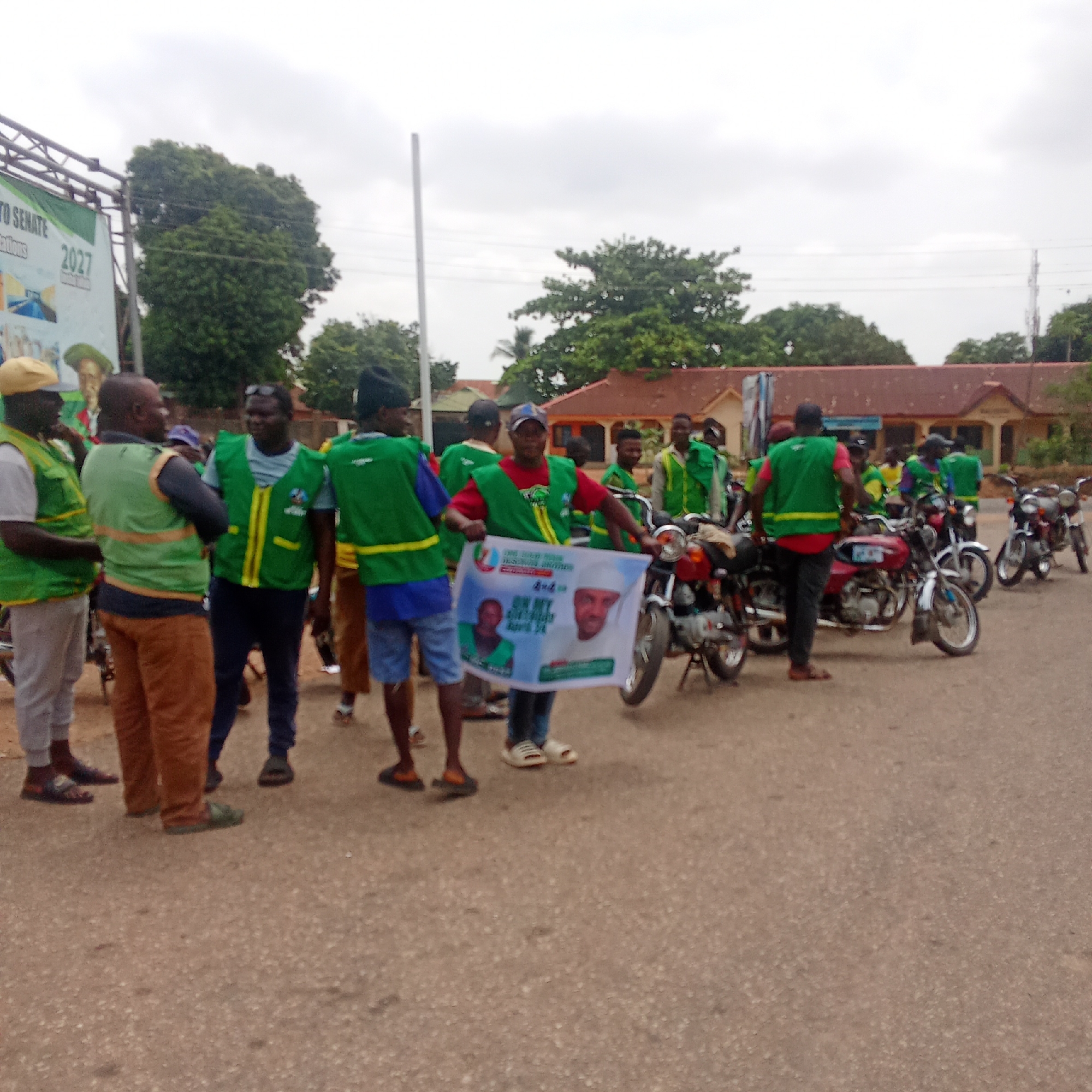 Vest-wearing group gathers with motorcycles, poster in Akwanga