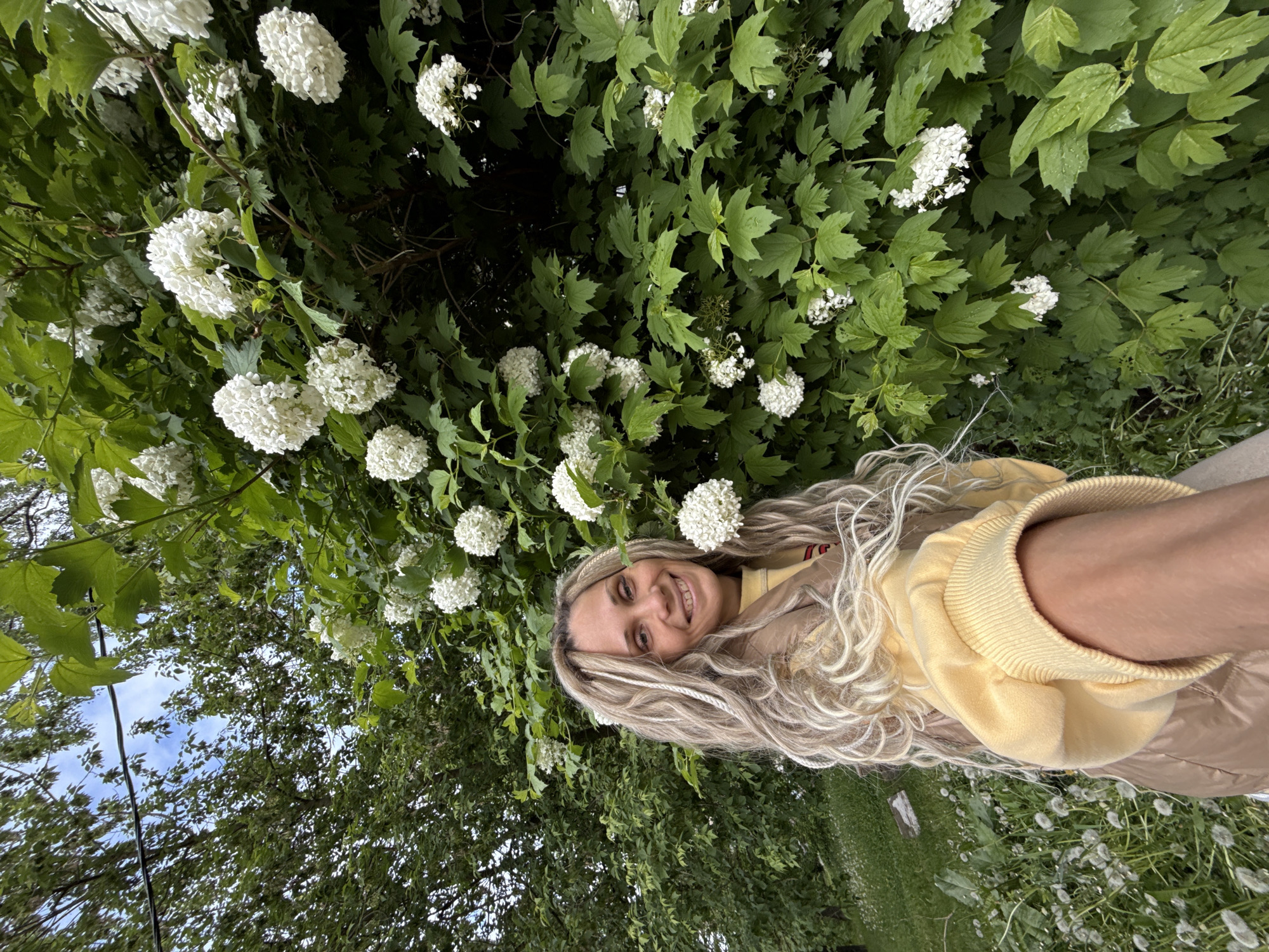 Woman in yellow sweatshirt photographed outdoors with flowering bushes