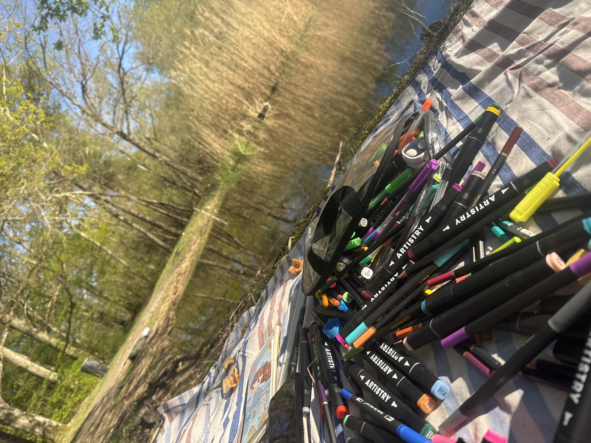 Artist with markers sits on blanket by water in Combreux