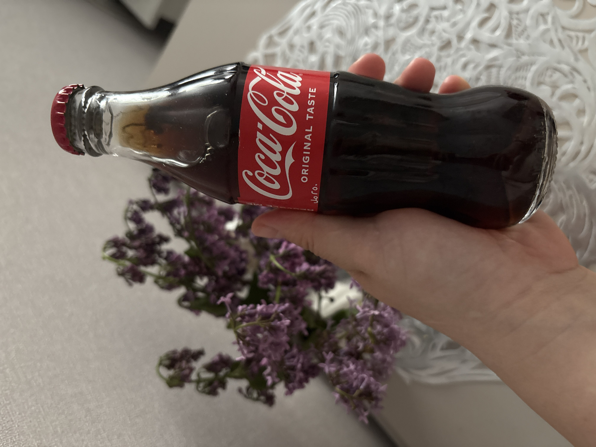 Person holds tilted Coca-Cola bottle above purple flowers