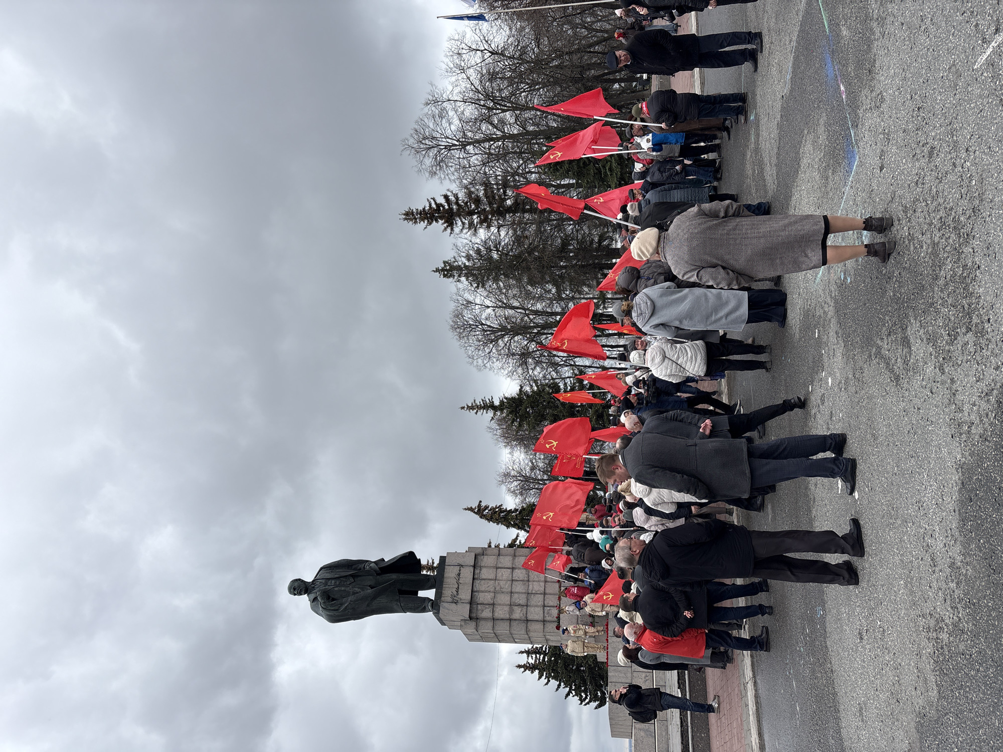 Elderly crowd gathers at Lenin statue with red flags