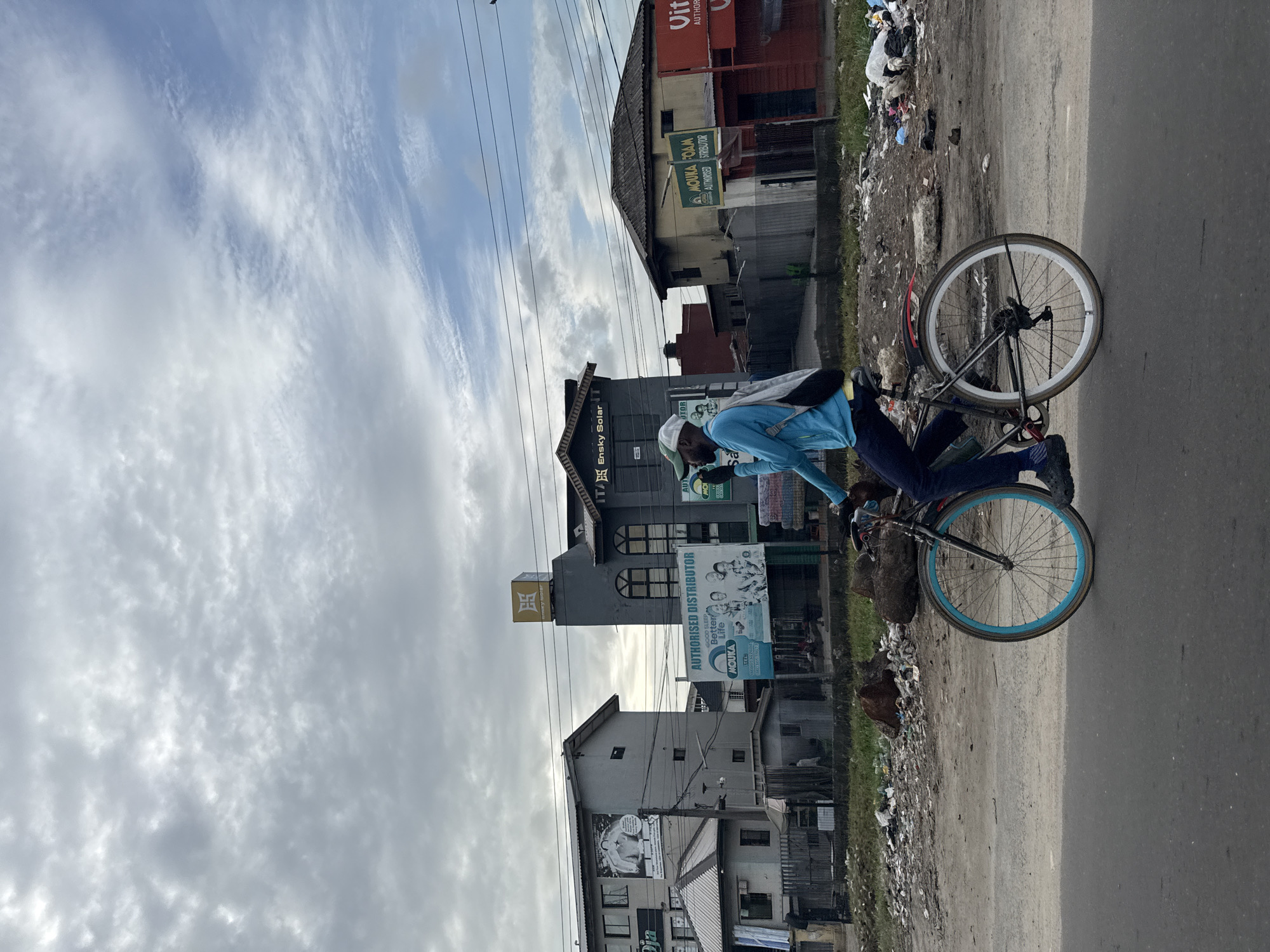 Cyclist rides blue-rimmed bicycle on debris-lined road in Nigeria