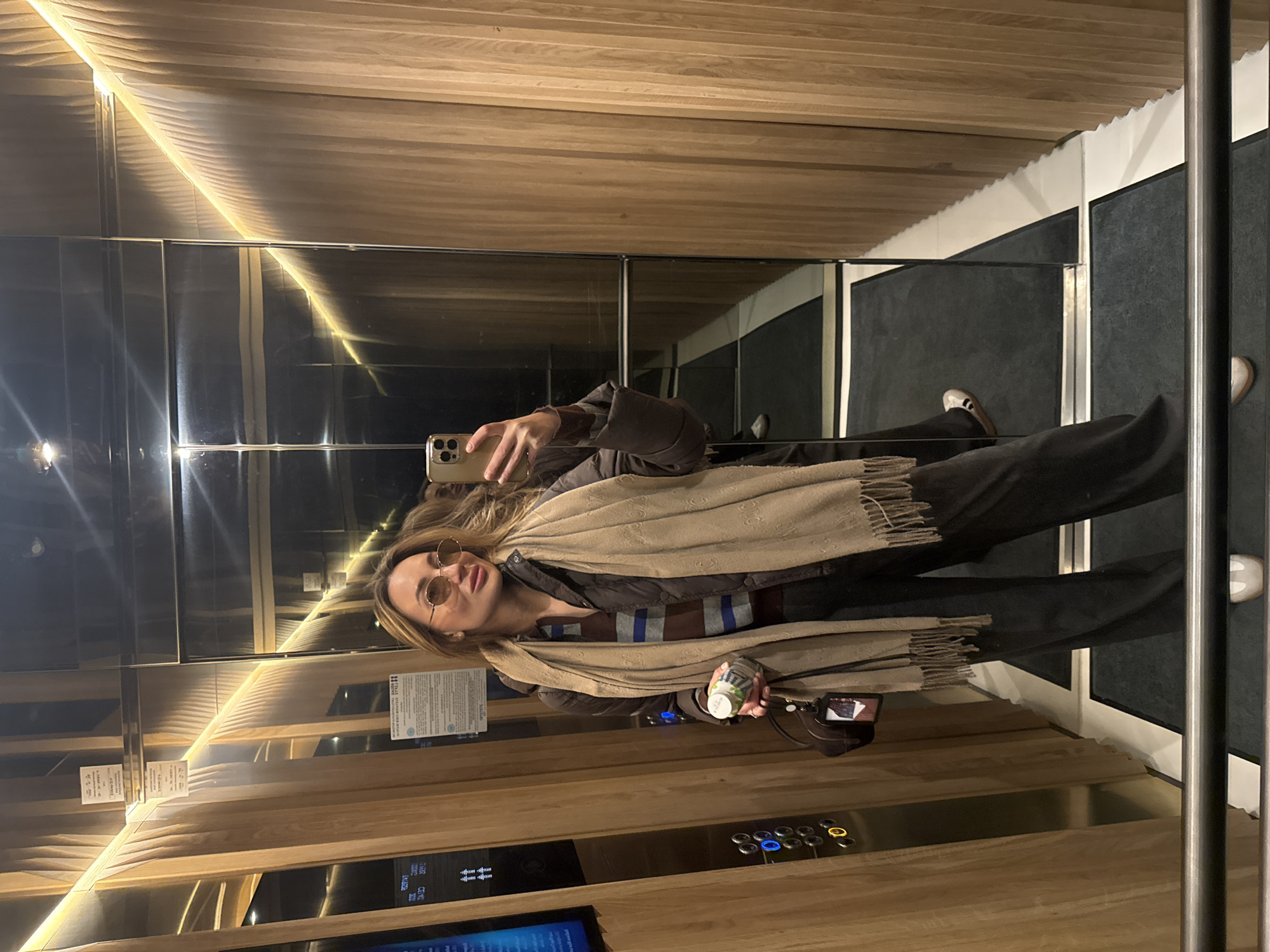 Woman takes selfie in wood-paneled elevator while holding bottle