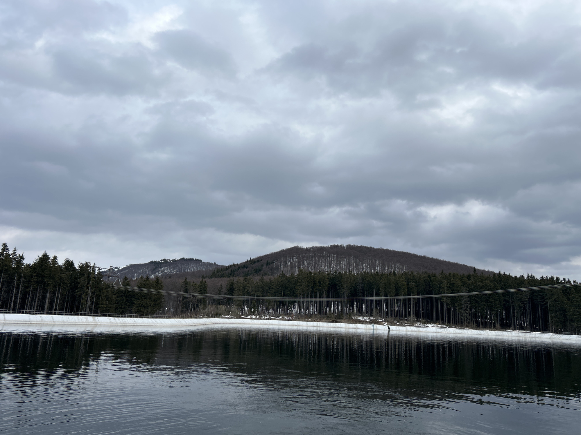 Calm lake scene documented amid forested hills, cloudy skies