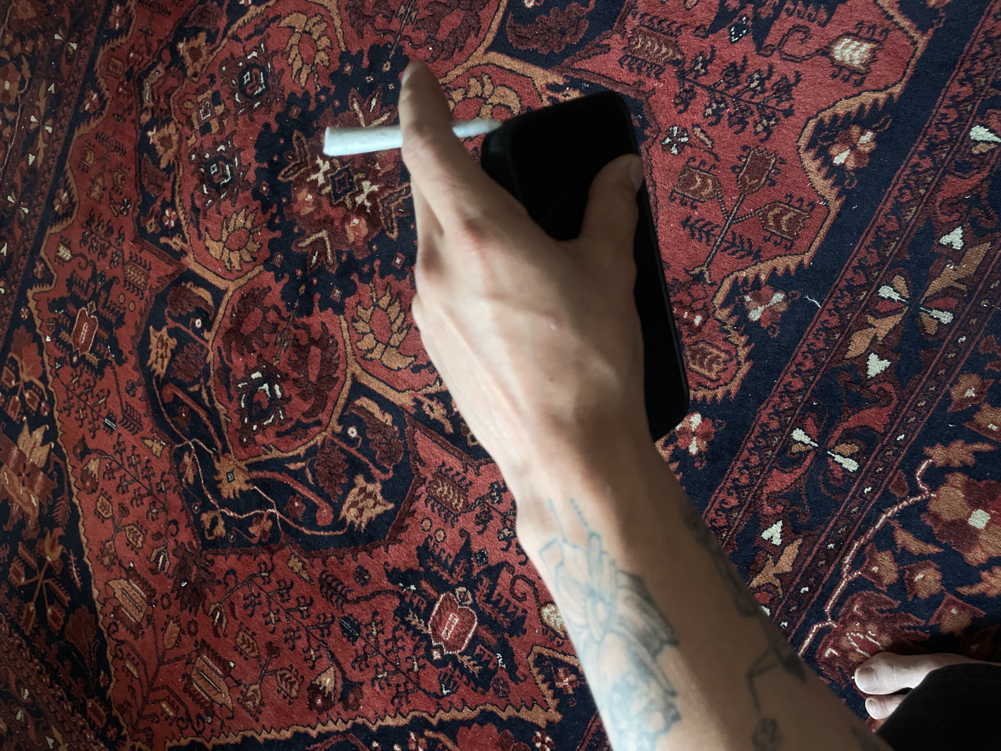 Tattooed hand holds cigarette and phone over patterned rug